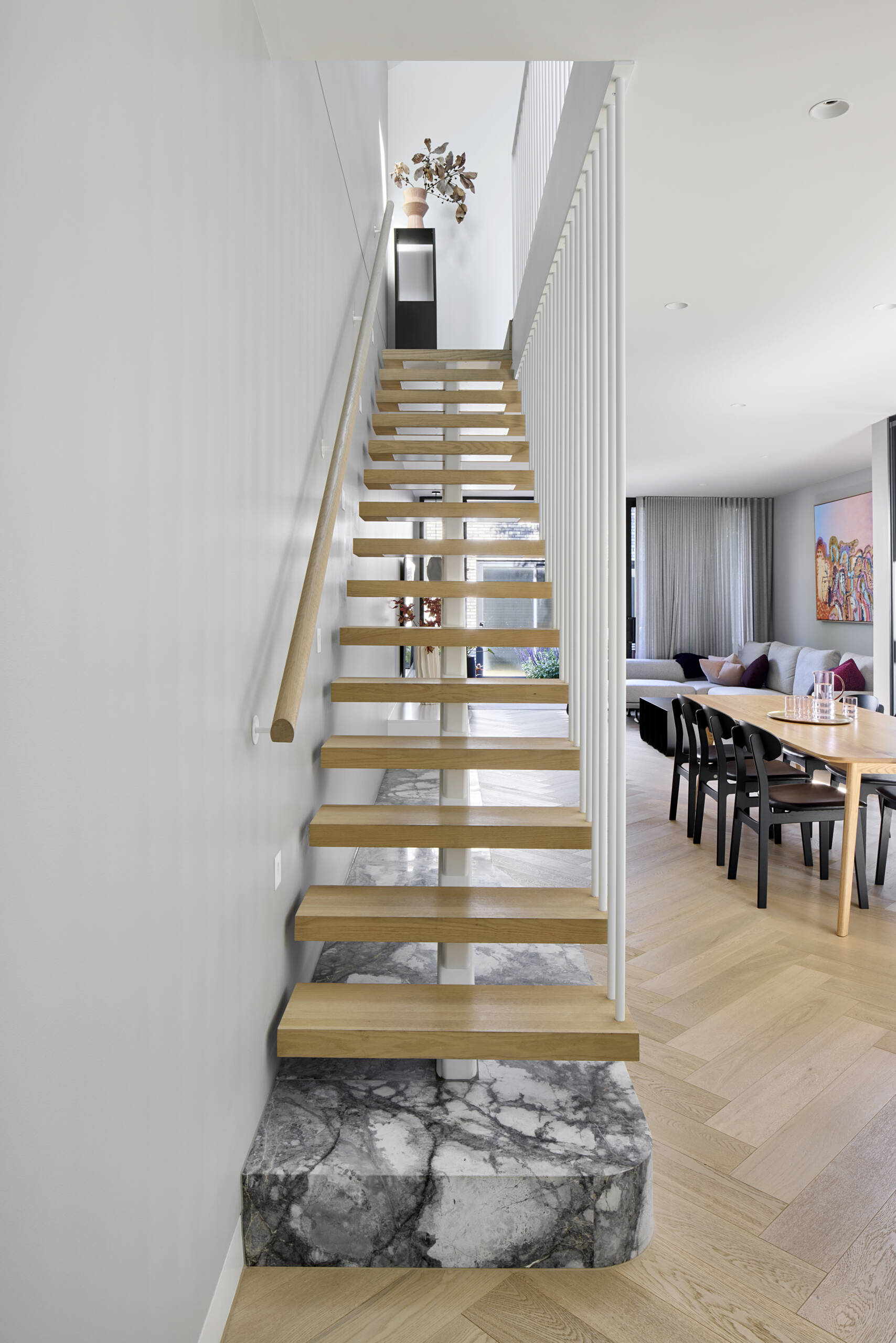 The staircase sits as a quiet feature, with pale wood treads floating above a marble platform. Slim white balusters keep the structure light. Grey walls and herringbone floors create a soft backdrop for simple pottery and branches that mark the transition between levels.