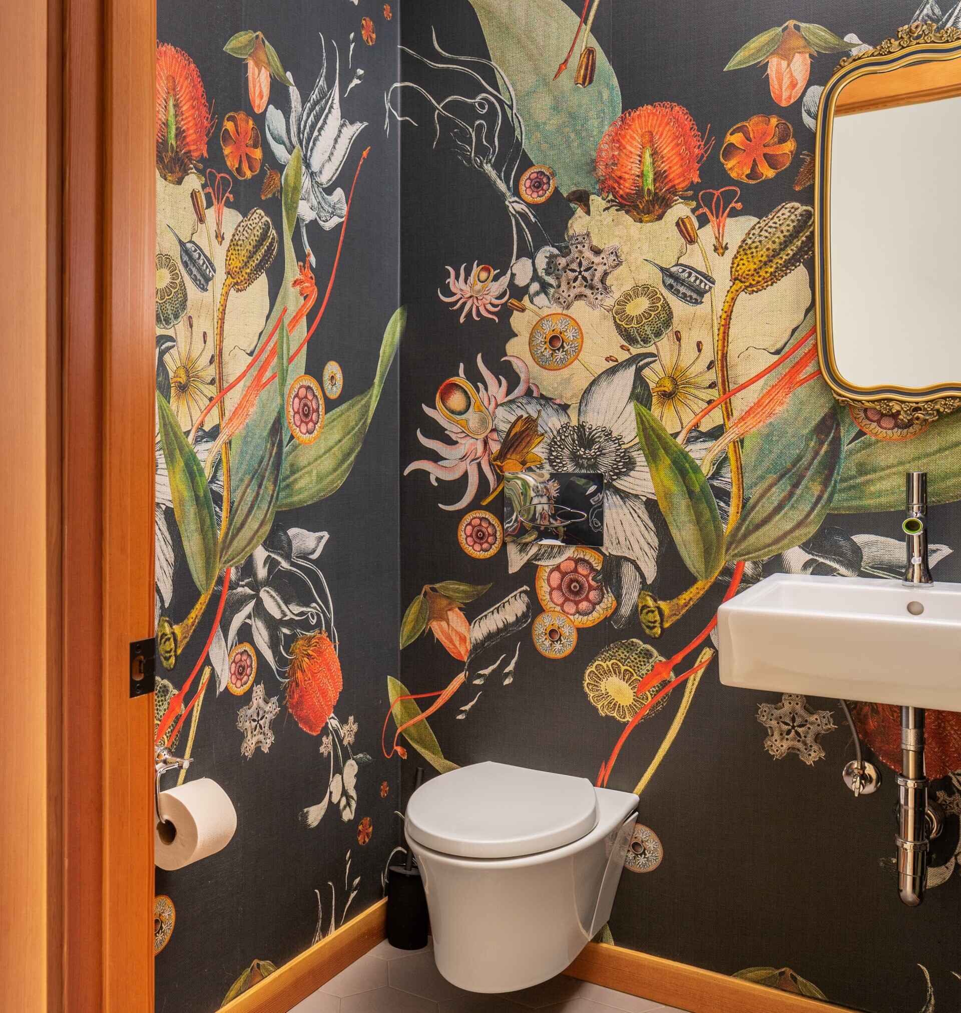 El tocador incorpora audazmente un llamativo papel tapiz botánico, llenando el pequeño espacio de color y movimiento. Un sencillo lavabo de pared y un sutil espejo enmarcado aportan equilibrio a la habitación.