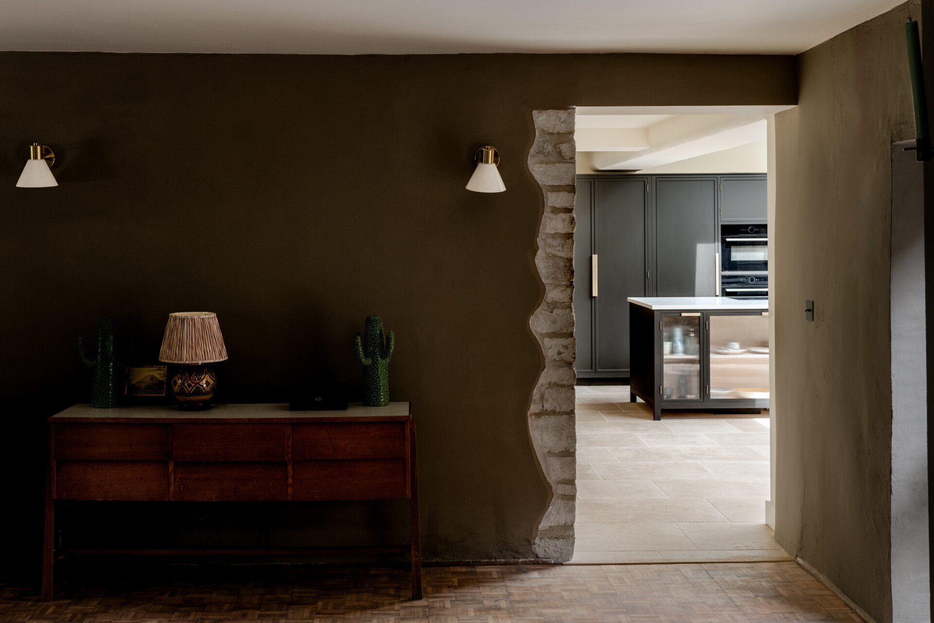 A character filled living room is anchored by a historic stone fireplace that's paired with a wood burning stove and mid century furniture. Deep wall tones, velvet upholstery, and natural textures create a warm, layered atmosphere that feels timeless and modern at once.