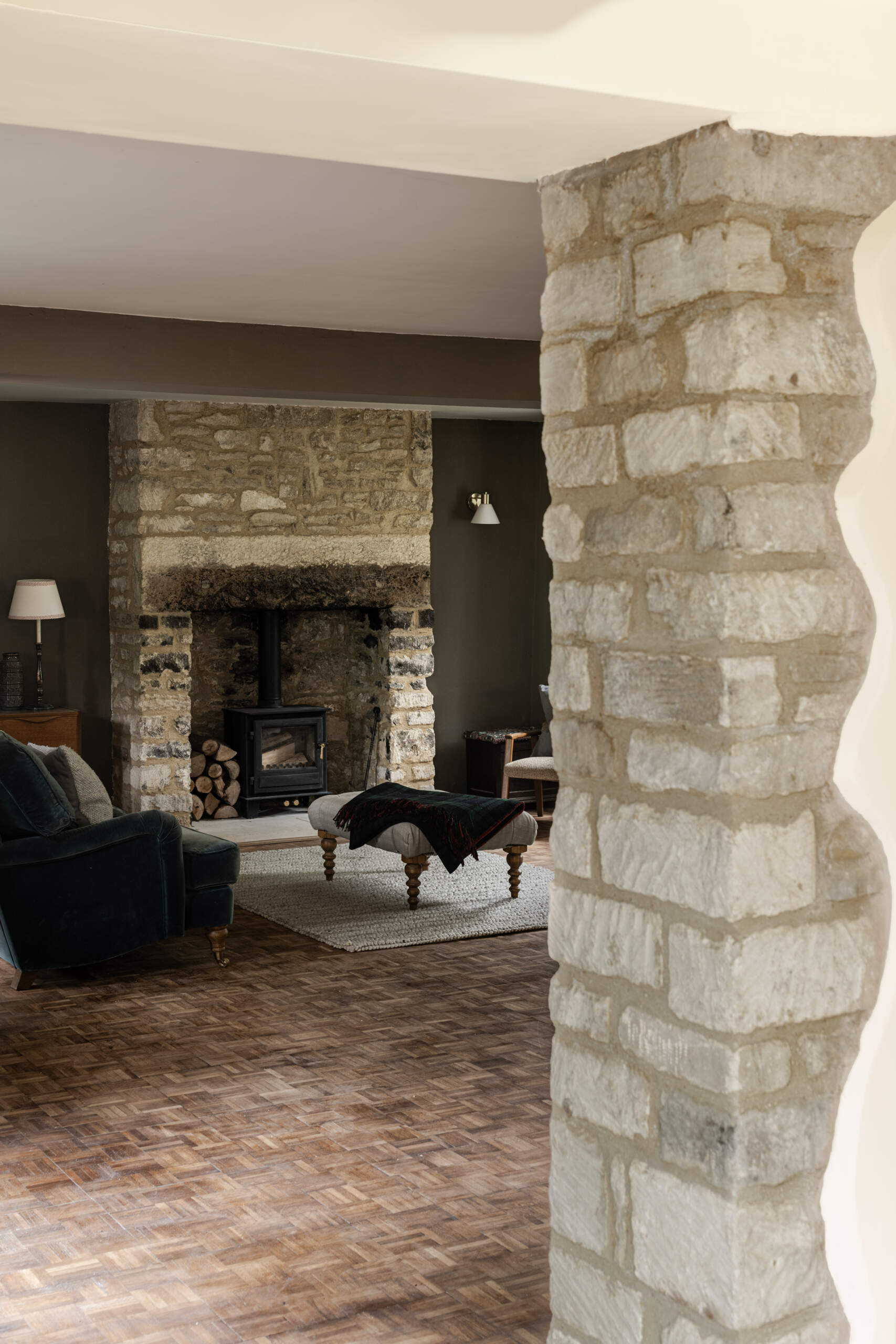 A character filled living room is anchored by a historic stone fireplace that's paired with a wood burning stove and mid century furniture. Deep wall tones, velvet upholstery, and natural textures create a warm, layered atmosphere that feels timeless and modern at once.