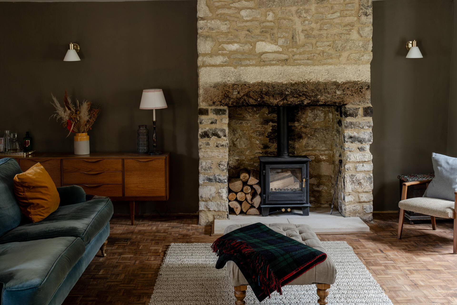 A character filled living room is anchored by a historic stone fireplace that's paired with a wood burning stove and mid century furniture. Deep wall tones, velvet upholstery, and natural textures create a warm, layered atmosphere that feels timeless and modern at once.