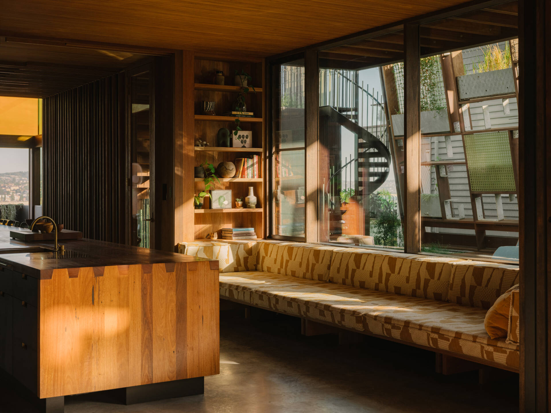 A long daybed sits within the kitchen area, offering a quiet spot to pause. It frames views of the pool and back yard, letting anyone resting there feel connected to the flow of the home. Soft light filtered through the plant canopy and stained glass creates a calm, sun-dappled space perfect for reading, lounging, or simply watching life move around the pool.