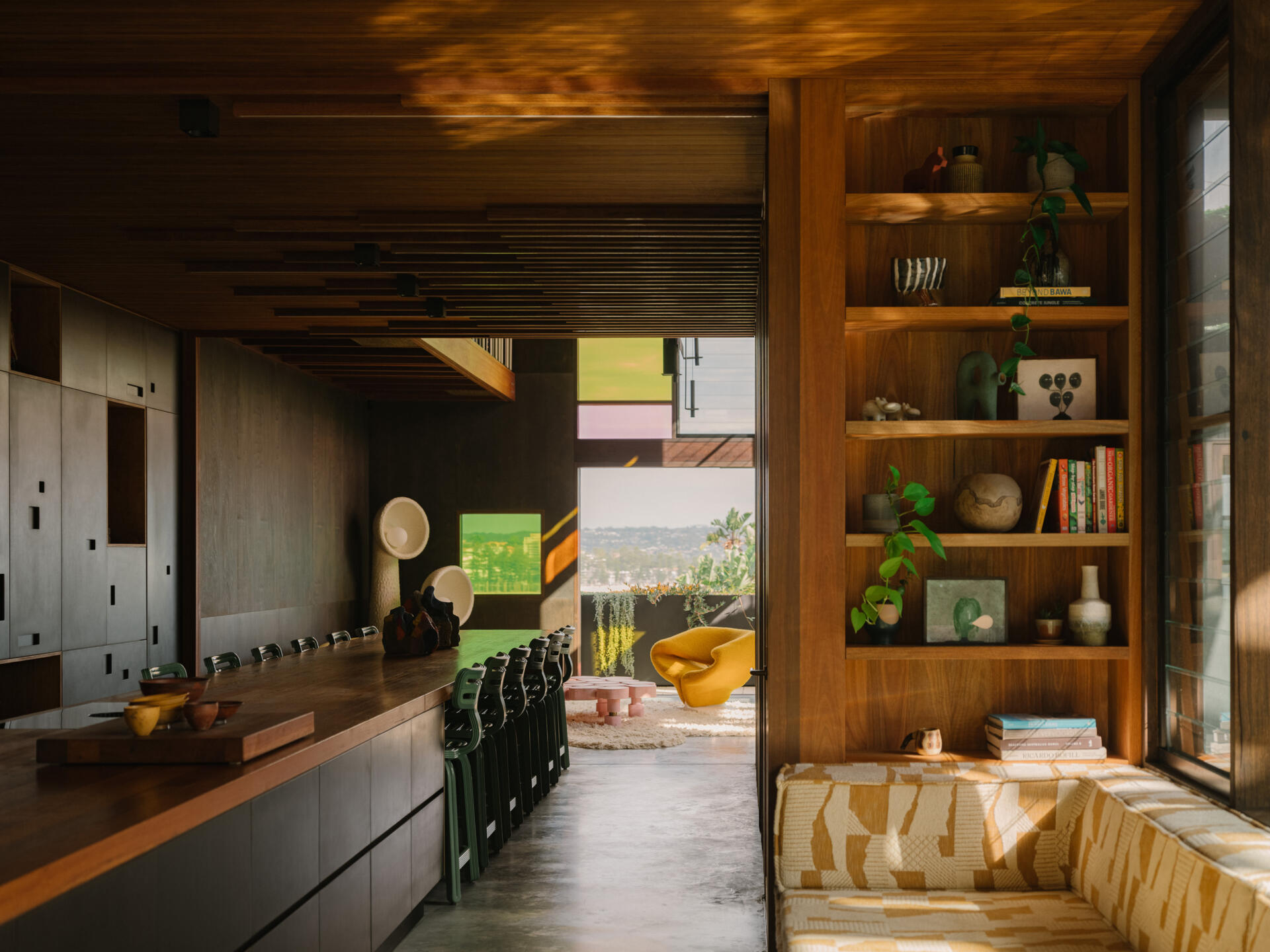 A long daybed sits within the kitchen area, offering a quiet spot to pause. It frames views of the pool and back yard, letting anyone resting there feel connected to the flow of the home. Soft light filtered through the plant canopy and stained glass creates a calm, sun-dappled space perfect for reading, lounging, or simply watching life move around the pool.