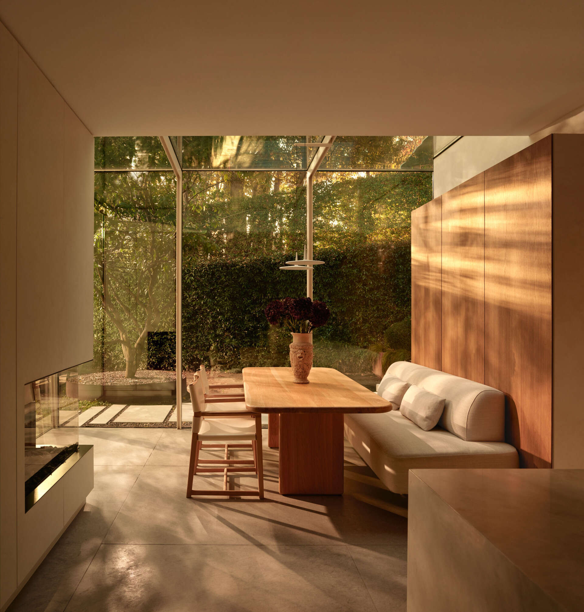 A casual dining nook has stripes of natural light highlighting warm wood paneling and a creamy banquette. The wood table and pared-back chairs maintain a relaxed atmosphere, while floor-to-ceiling glass connects the space to the garden.