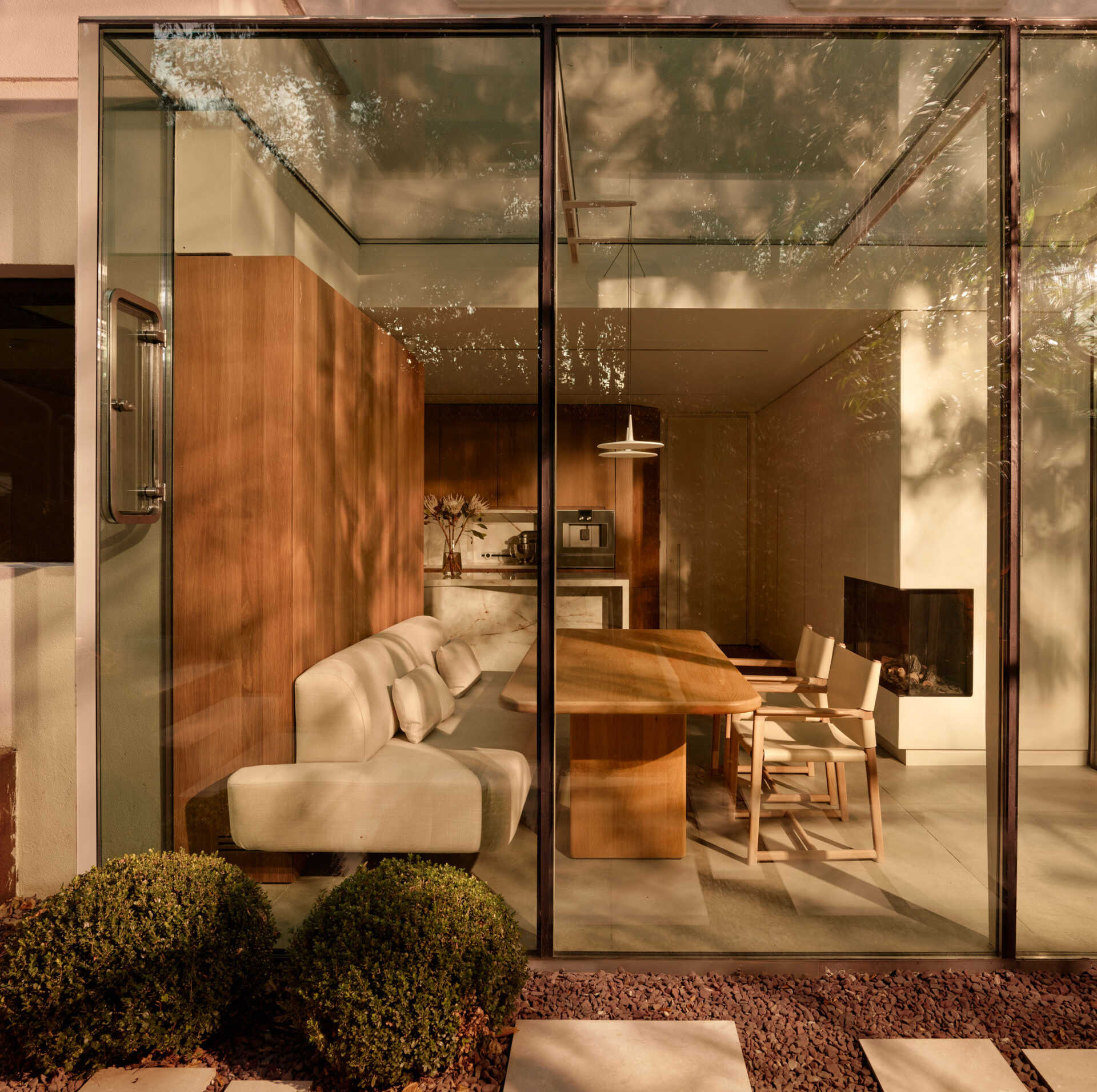 A casual dining nook has stripes of natural light highlighting warm wood paneling and a creamy banquette. The wood table and pared-back chairs maintain a relaxed atmosphere, while floor-to-ceiling glass connects the space to the garden.