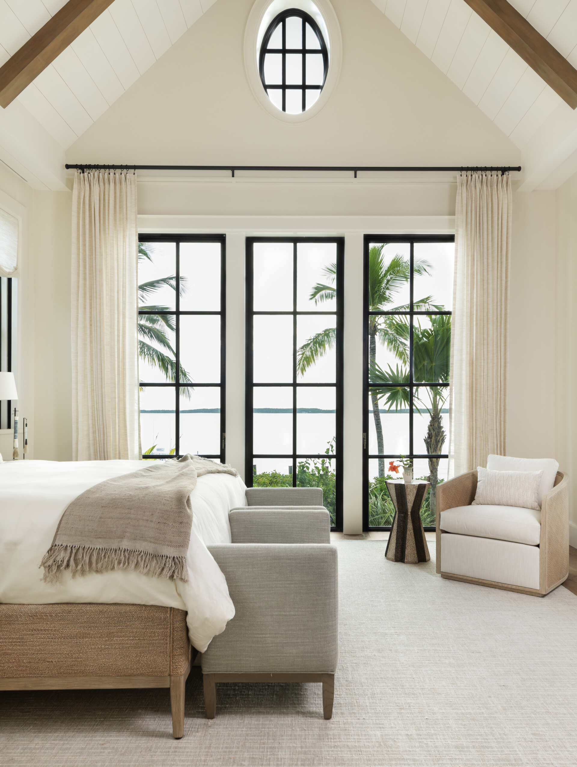 Calm island bedroom layered with soft whites, warm wood, and natural textures, where black framed doors and windows frame palm and ocean views like living artwork.