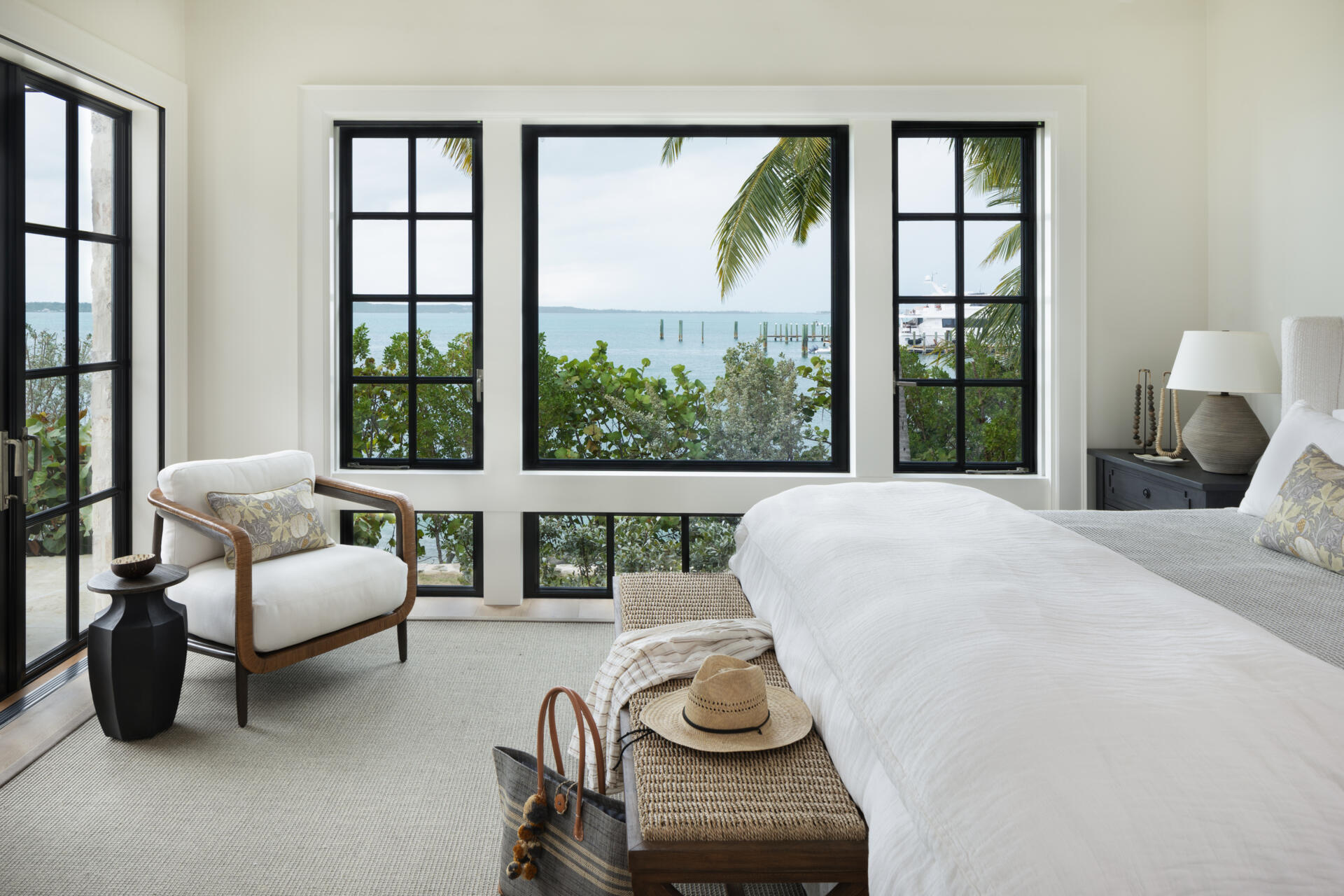 Calm island bedroom layered with soft whites, warm wood, and natural textures, where black framed doors and windows frame palm and ocean views like living artwork.