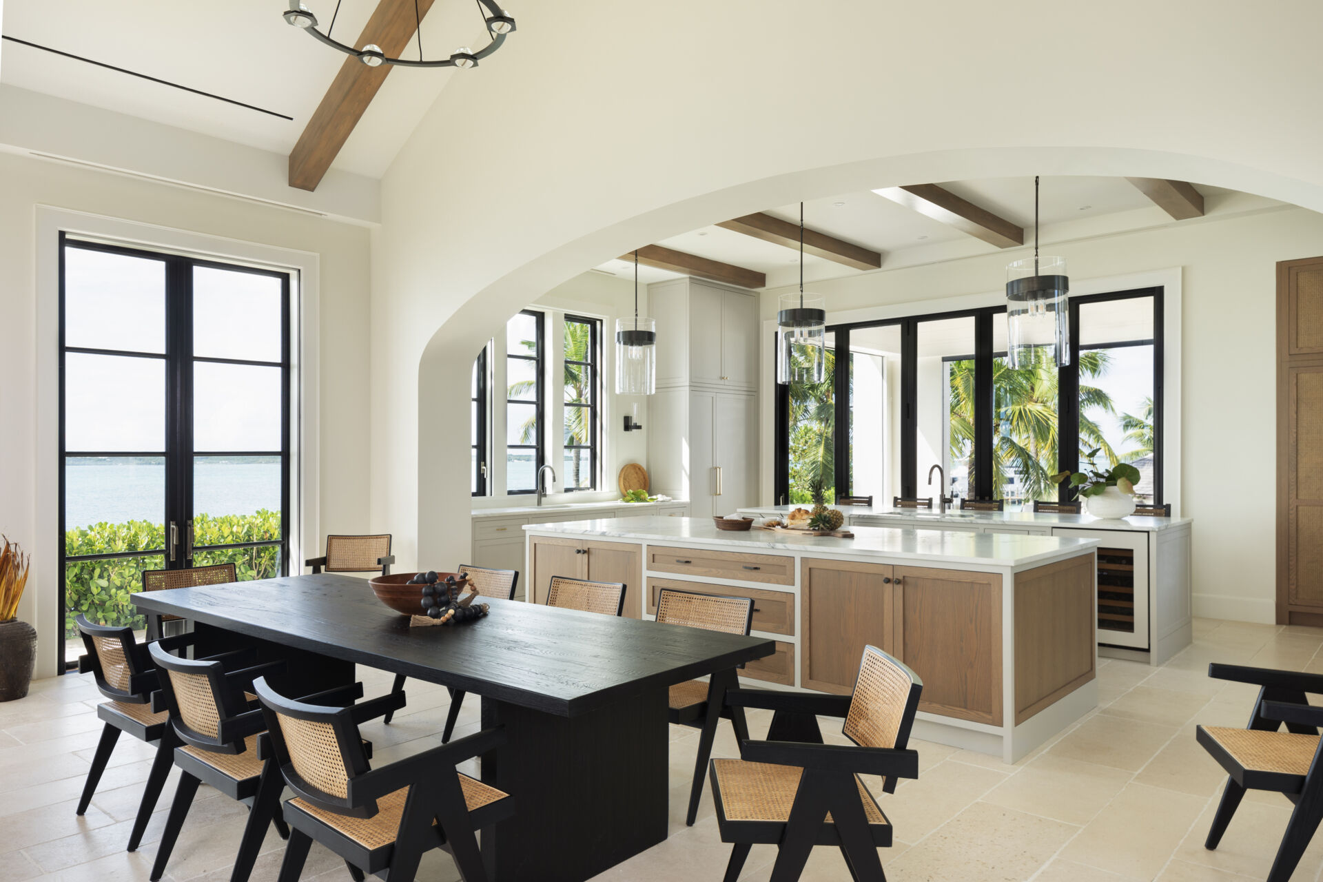 Dining area that flows seamlessly between the living space and kitchen, grounded by pale stone floors, woven seating, and views framed by black windows for long, social meals.