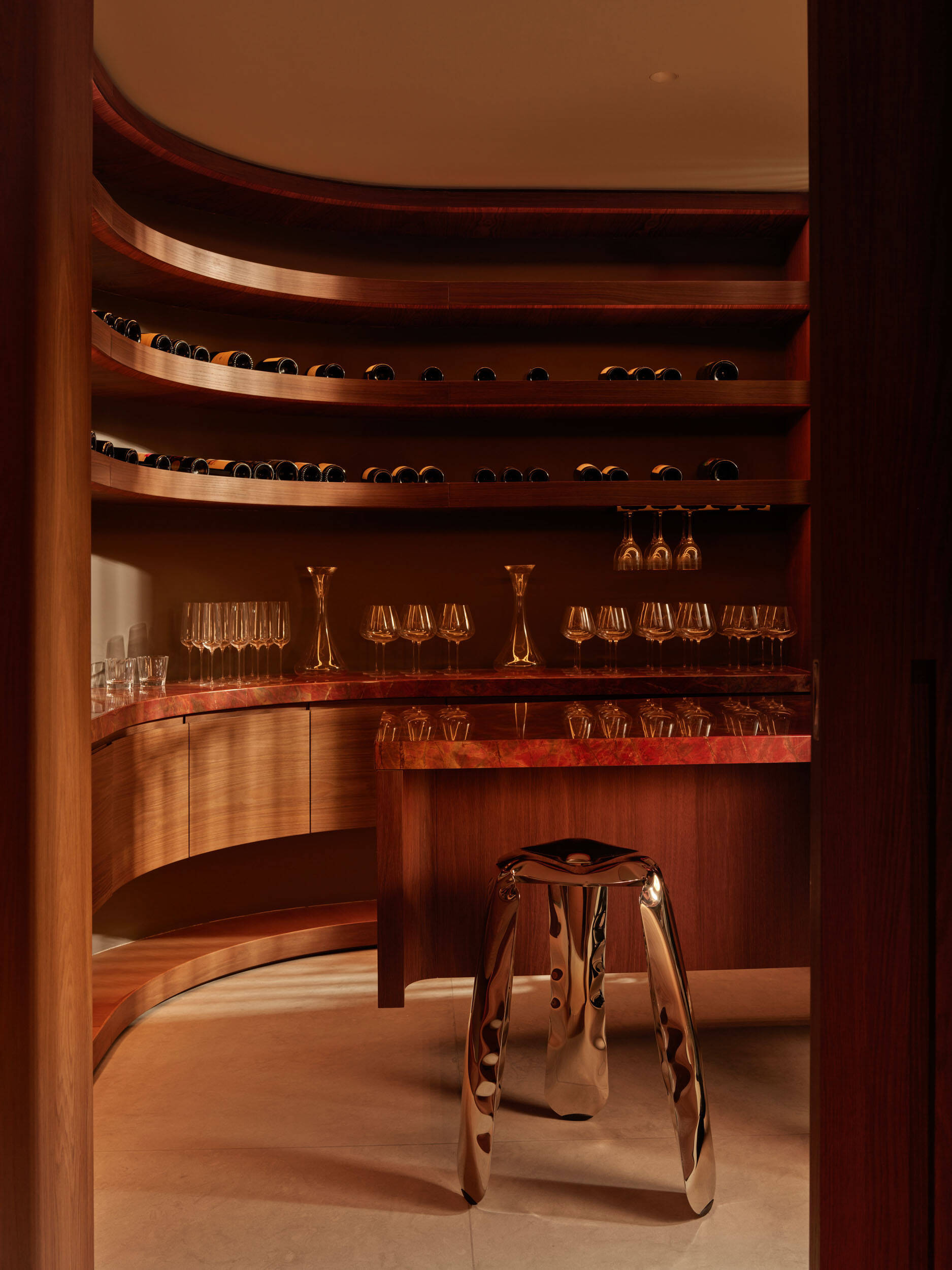 A curved bar nook feels like a hidden lounge, wrapped in rich wood shelving with rows of wine bottles. Amber lighting warms the reddish stone countertop. Rounded cabinetry softens the room’s edges, while a mirror-finish sculptural stool adds a modern twist.