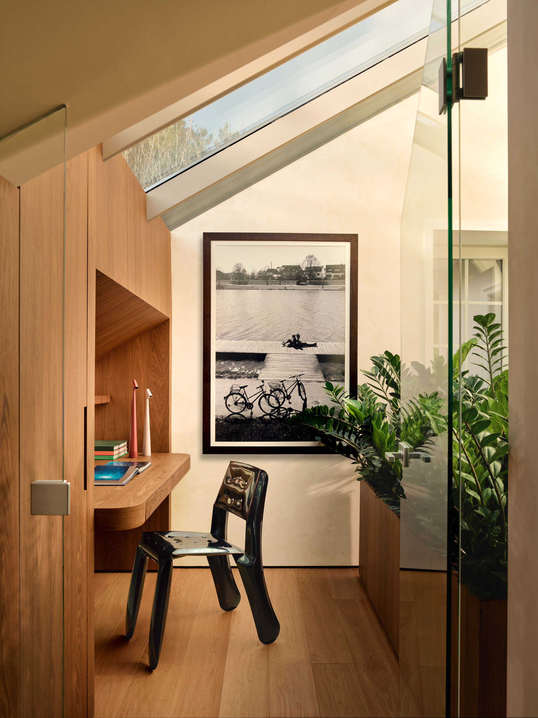 The home office feels bright and focused, with warm wood built-ins keeping everything organized. A skylight brings natural light and greenery act as a calming backdrop. A glossy sculptural chair adds personality, while a large black-and-white photograph introduces a quiet, lived-in feel.