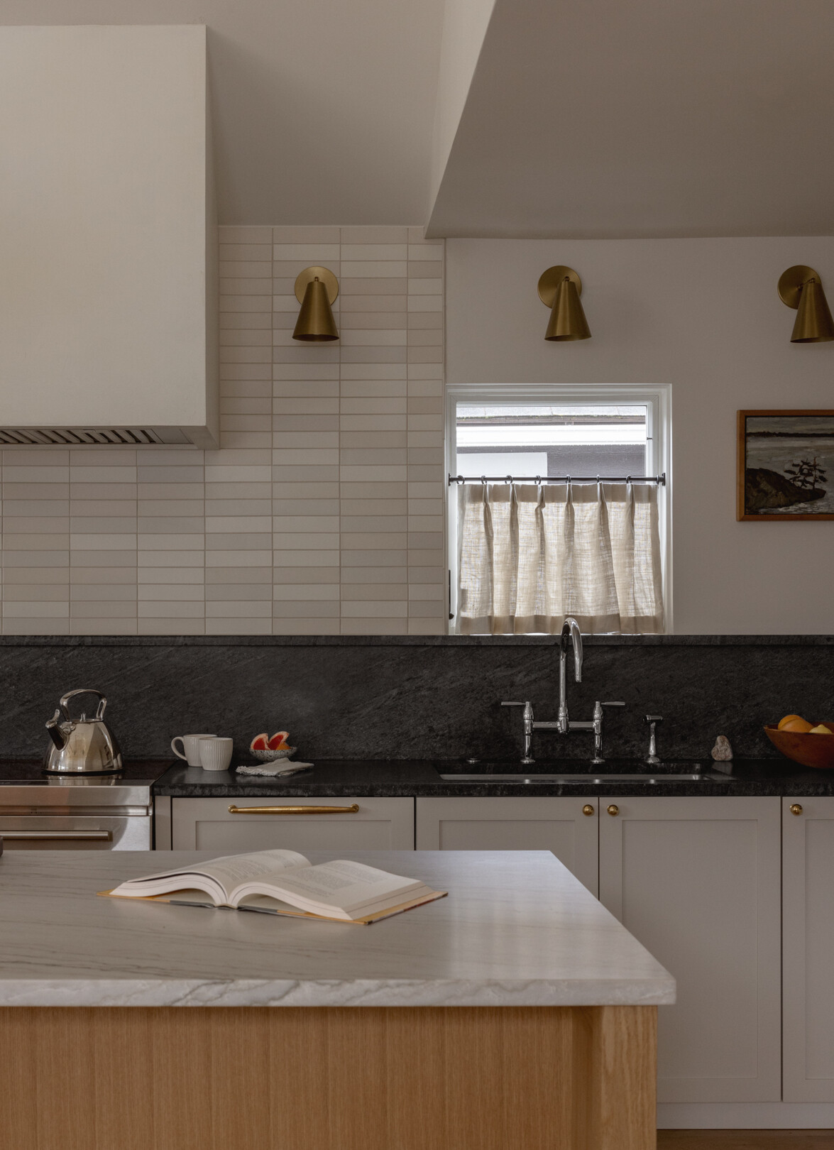 En esta cocina, gabinetes estilo Shaker pintados en tonos tranquilos y neutros se ubican junto a una isla de roble blanco rematada con cuarcita perla marina, creando una pieza central cálida y relajante. Los mostradores perimetrales de esteatita añaden profundidad, mientras que una combinación de herrajes de latón, azulejos blancos cálidos y accesorios de cromo pulido aportan una variación sutil sin desorden visual.