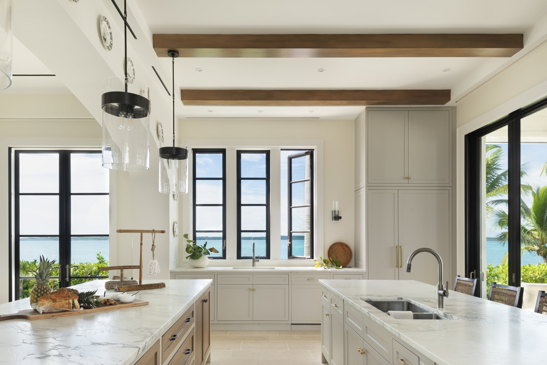 Bright Caribbean kitchen with dual islands, designed as the heart of the home, with pale cabinetry, timber ceiling beams, and large black framed windows that turn everyday moments into view-focused rituals.