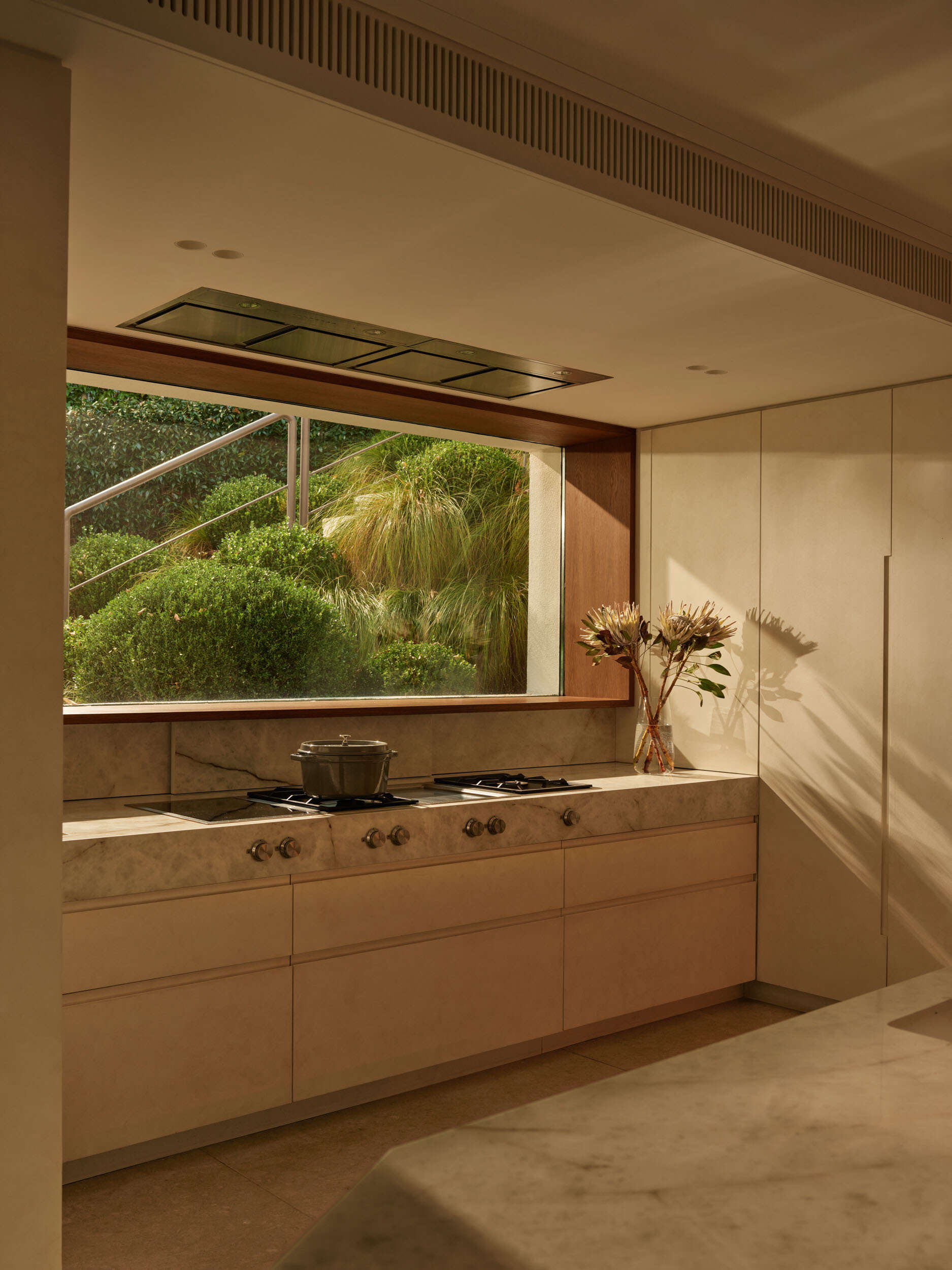 The kitchen feels serene, grounded in natural flat-panel cabinetry and a soft stone countertop. A wide picture window frames lush greenery, bringing nature into the workspace. Warm wood trim adds depth, while a simple vase of blooms introduces an organic note.