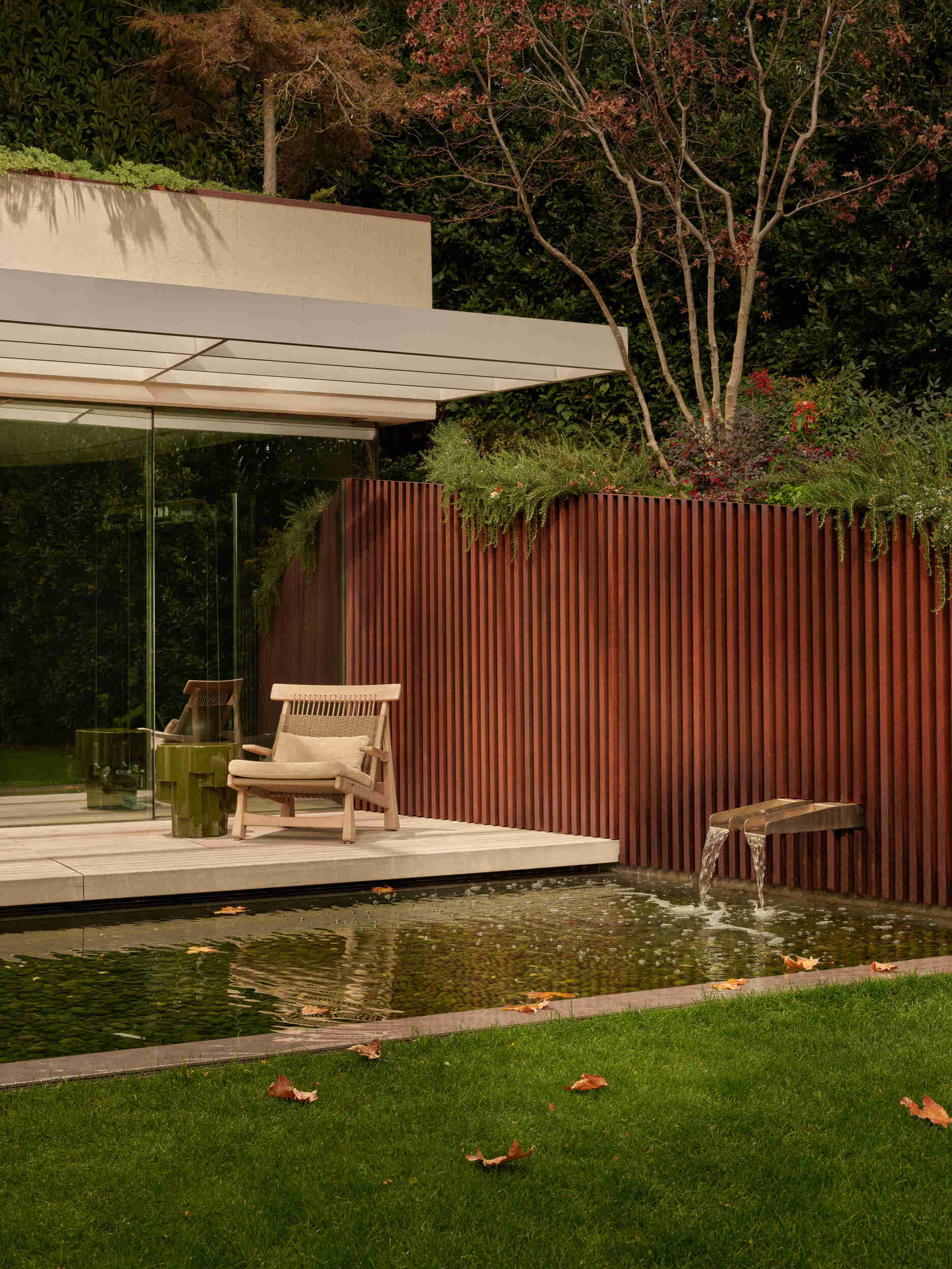 Outside, a sleek reflecting pool reveals a pebbled bottom like a natural mosaic. Pale deck boards curve around the water, while a vertical screen provides privacy. A lounge chair and sculptural side table complete this calm, uncluttered space ideal for quiet afternoons.