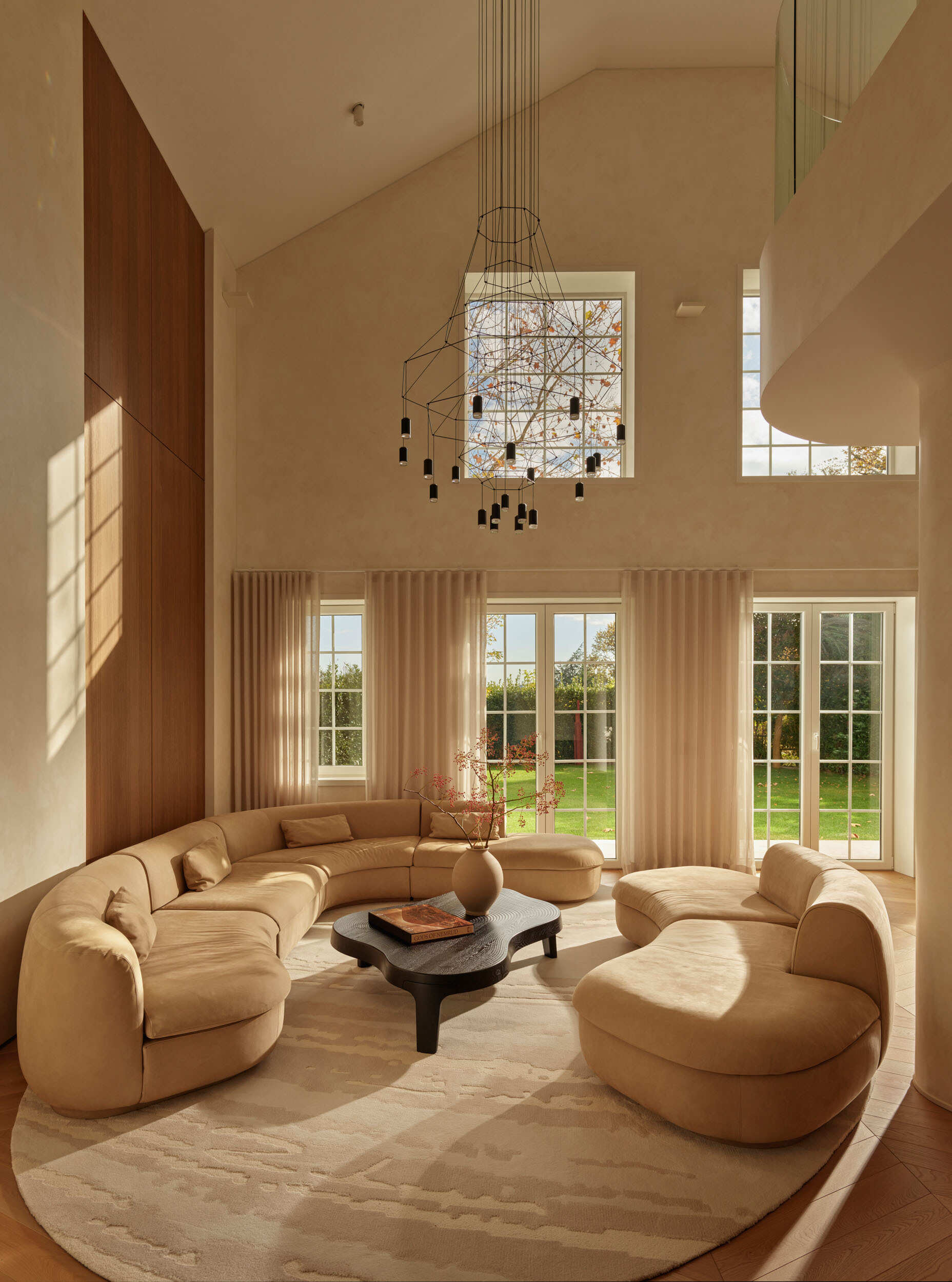 The living room is bright and airy, with cream sofas arranged in a conversation circle atop a soft patterned rug. Tall French doors let sunlight flood the space, bringing the garden indoors. A sculptural black coffee table adds a modern edge, while the high ceiling and delicate pendant fixture create a sense of openness.