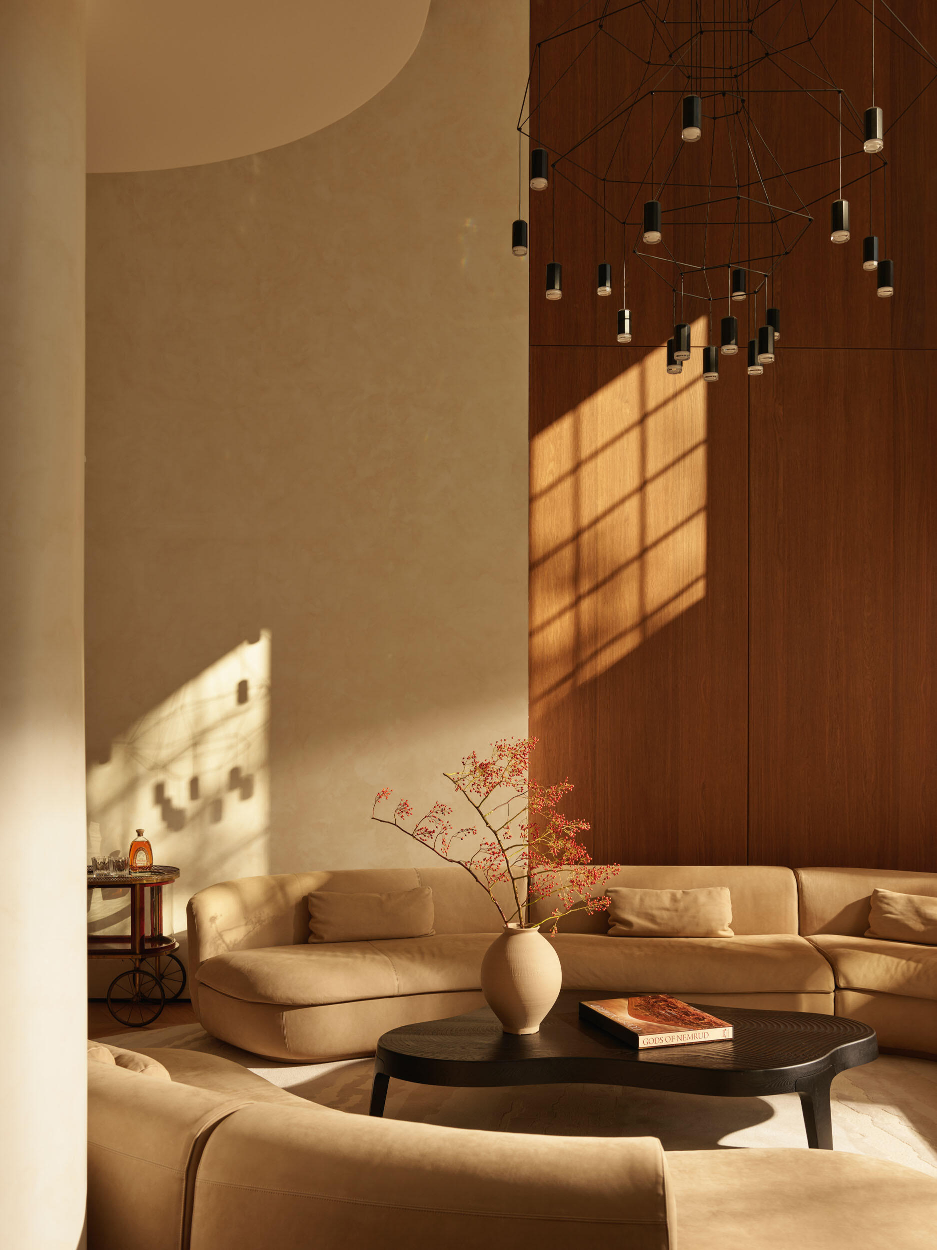 The living room is bright and airy, with cream sofas arranged in a conversation circle atop a soft patterned rug. Tall French doors let sunlight flood the space, bringing the garden indoors. A sculptural black coffee table adds a modern edge, while the high ceiling and delicate pendant fixture create a sense of openness.