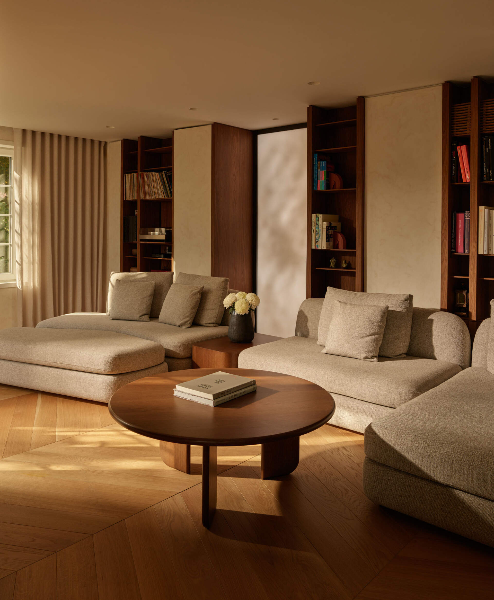 This secondary living area is soft and inviting, with oatmeal-toned seating and deep cushions for lounging. Wood floors add warmth, while built-in shelves keep the space tidy. Sunlight filters through full-height drapes, casting a gentle glow over the round wood coffee table.