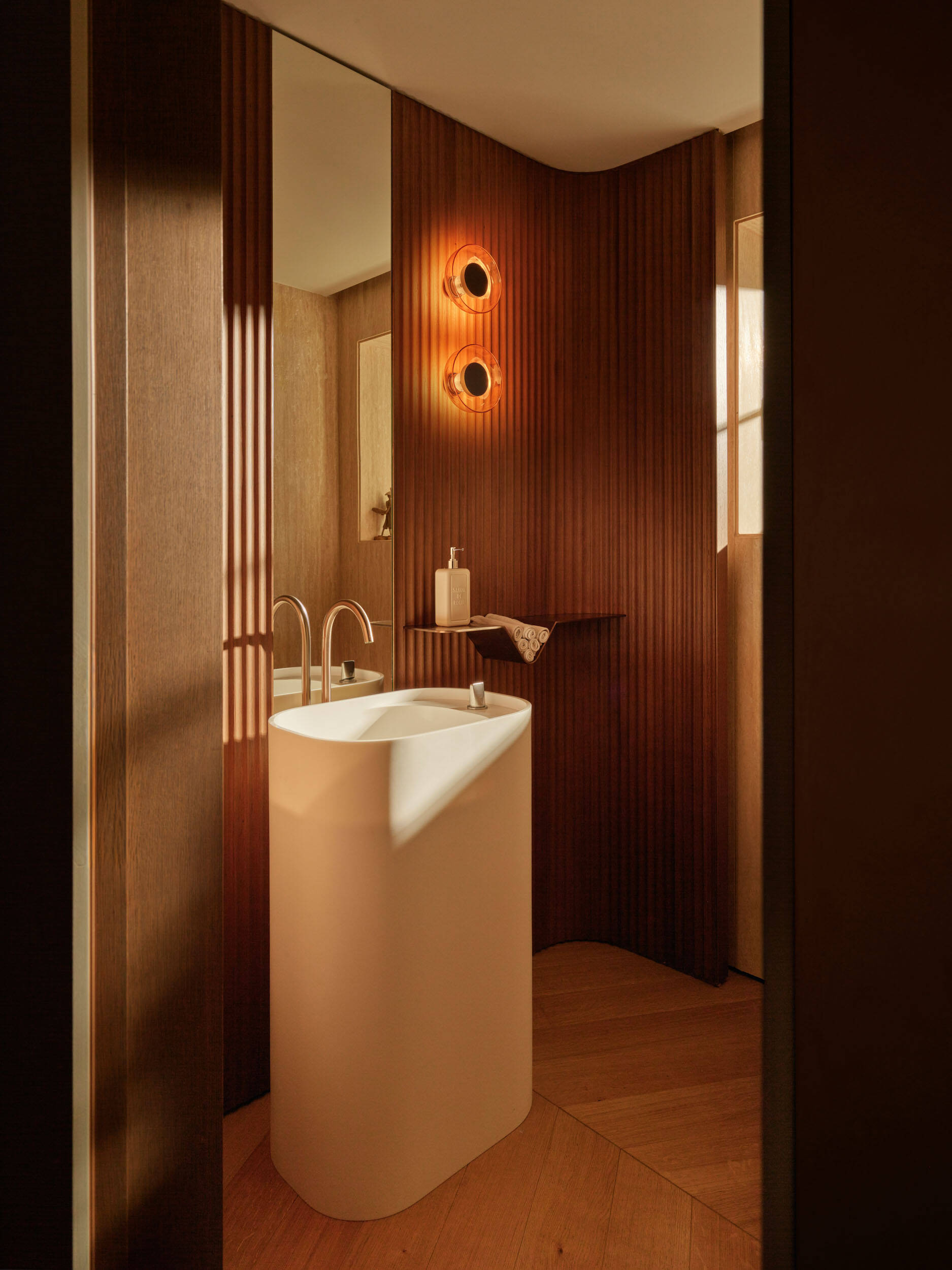 The powder room is a tiny jewel, wrapped in warm wood slats. A sculptural white pedestal sink keeps the footprint light, while chevron wood flooring softens the angles. Amber wall sconces cast a golden glow, and a slim shelf holds neatly rolled towels, keeping the space calm and intentional.