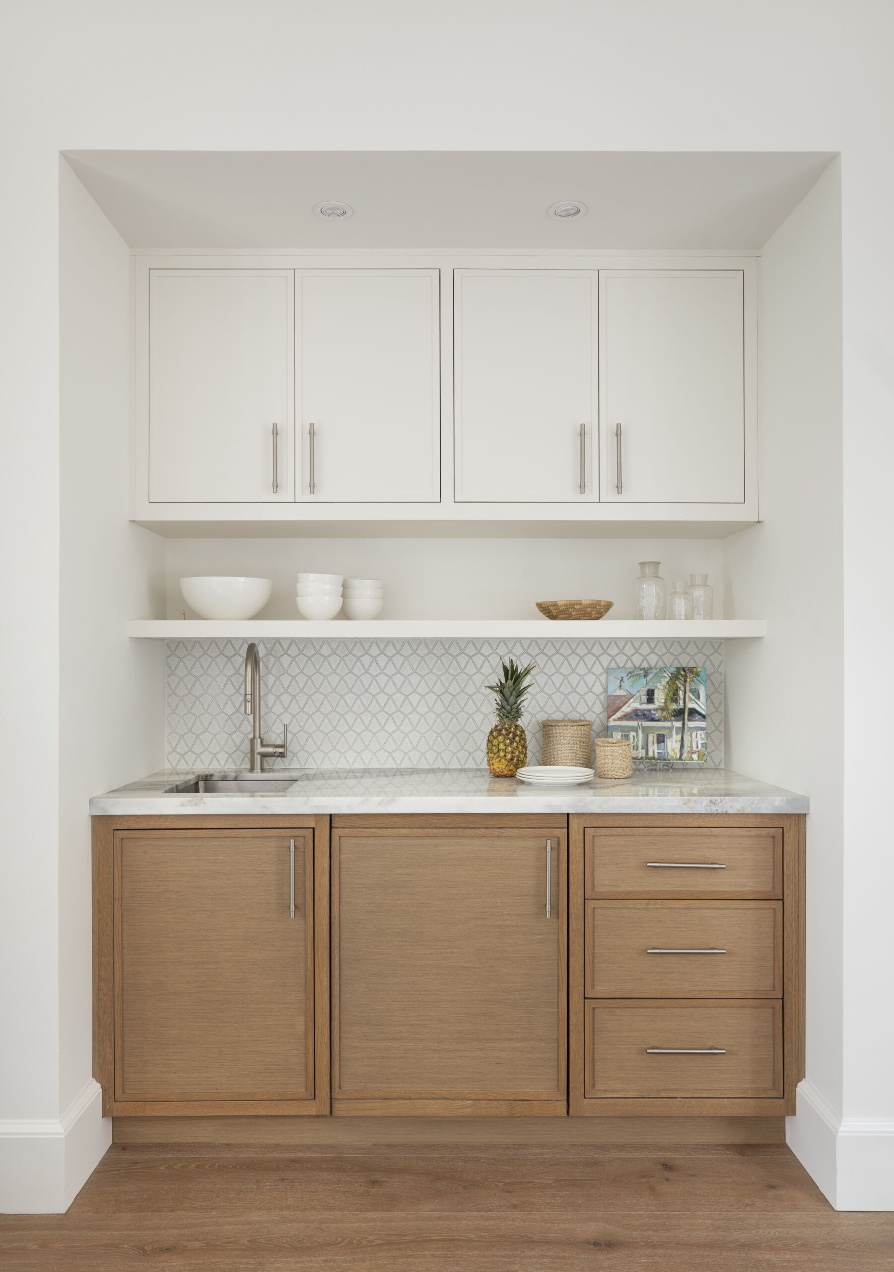 Compact wet bar that blends crisp white cabinetry with warm wood lowers, open shelving, and subtle patterning for easy, casual entertaining that feels effortless.