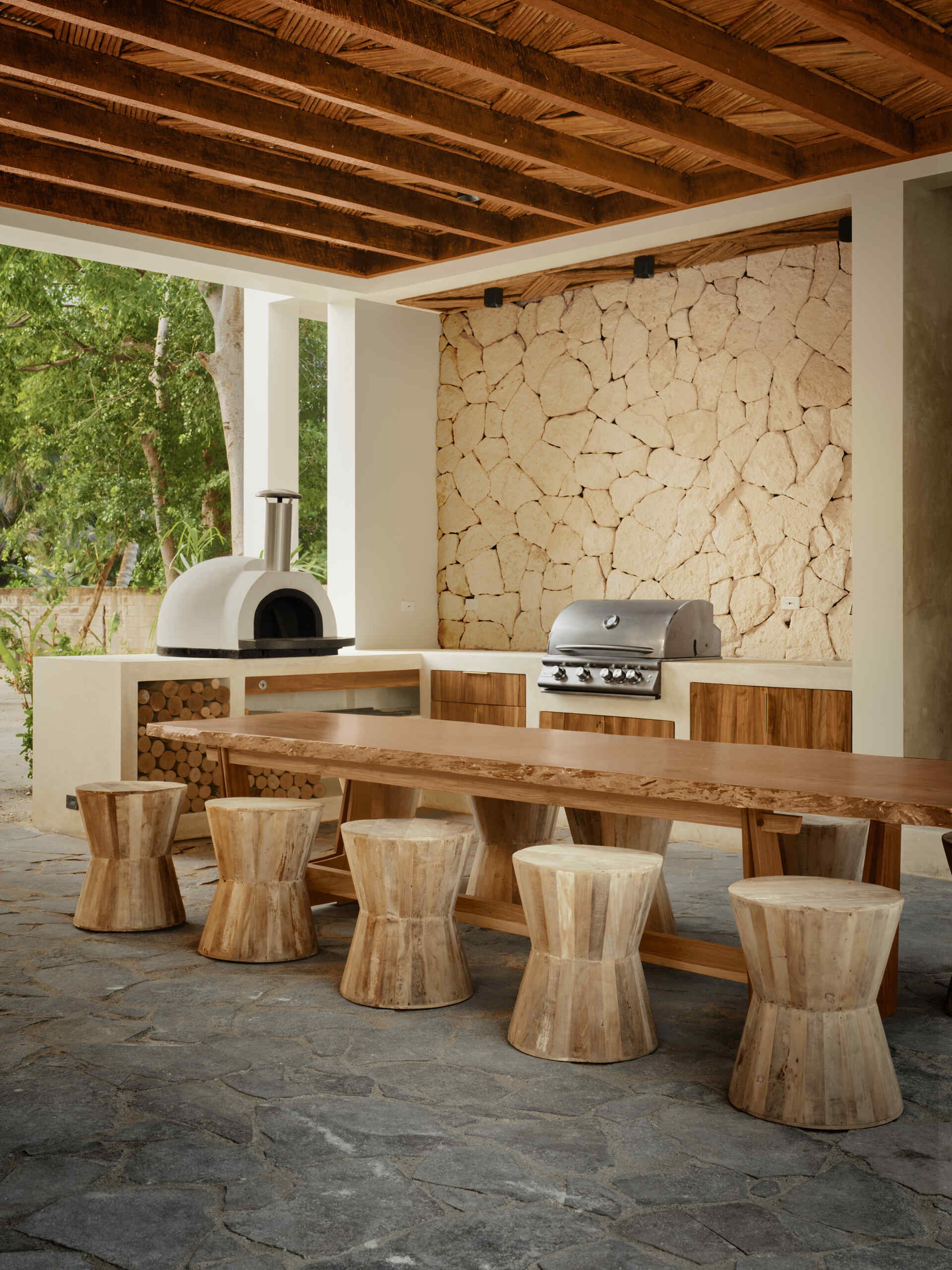 Outdoor cooking plays a central role in how the home functions. The outdoor kitchen includes a wood fired pizza oven, smoker, and grill, turning meals into shared events, while the dining area is furnished with an oversized wood table and stools.