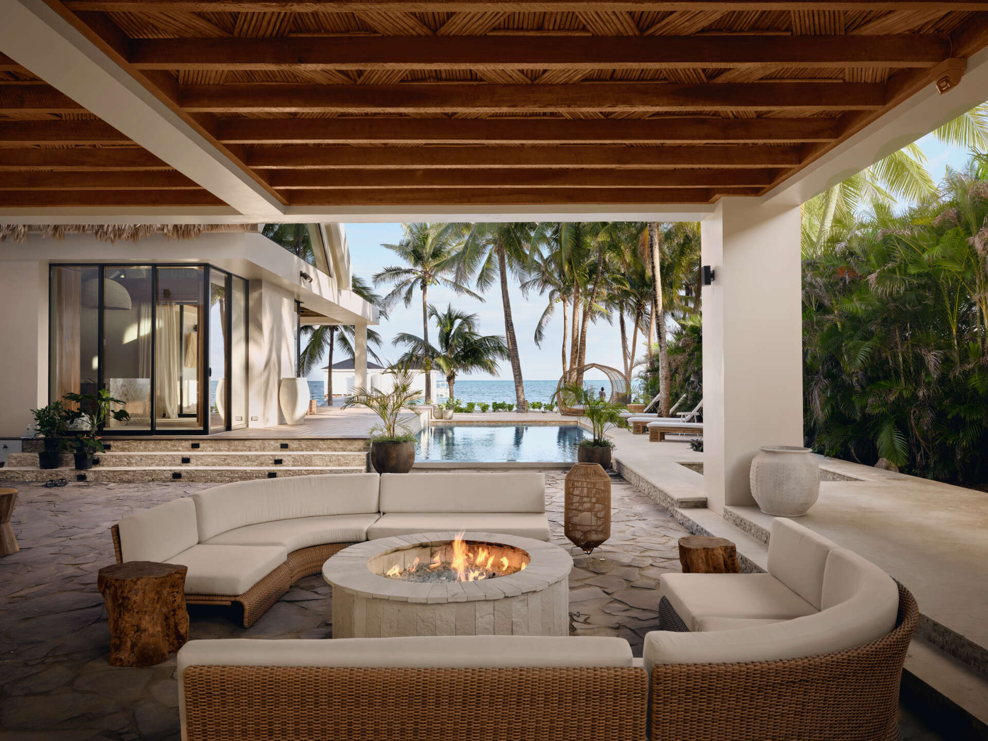 This outdoor lounge is arranged as a natural gathering point, sheltered beneath a wood lined ceiling and open on all sides to the landscape beyond. Curved seating wraps around a circular fire pit, creating a relaxed setting for conversation that continues into the evening. Stone paving grounds the space, while woven furniture and soft cushions add warmth and comfort.