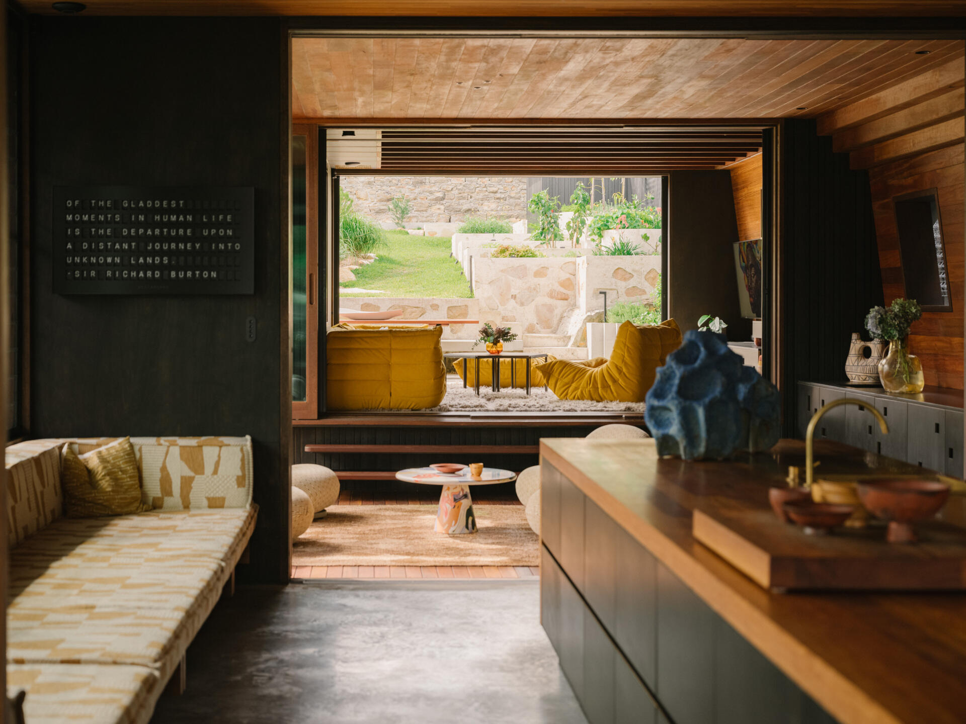 A long daybed sits within the kitchen area, offering a quiet spot to pause. It frames views of the pool and back yard, letting anyone resting there feel connected to the flow of the home. Soft light filtered through the plant canopy and stained glass creates a calm, sun-dappled space perfect for reading, lounging, or simply watching life move around the pool.