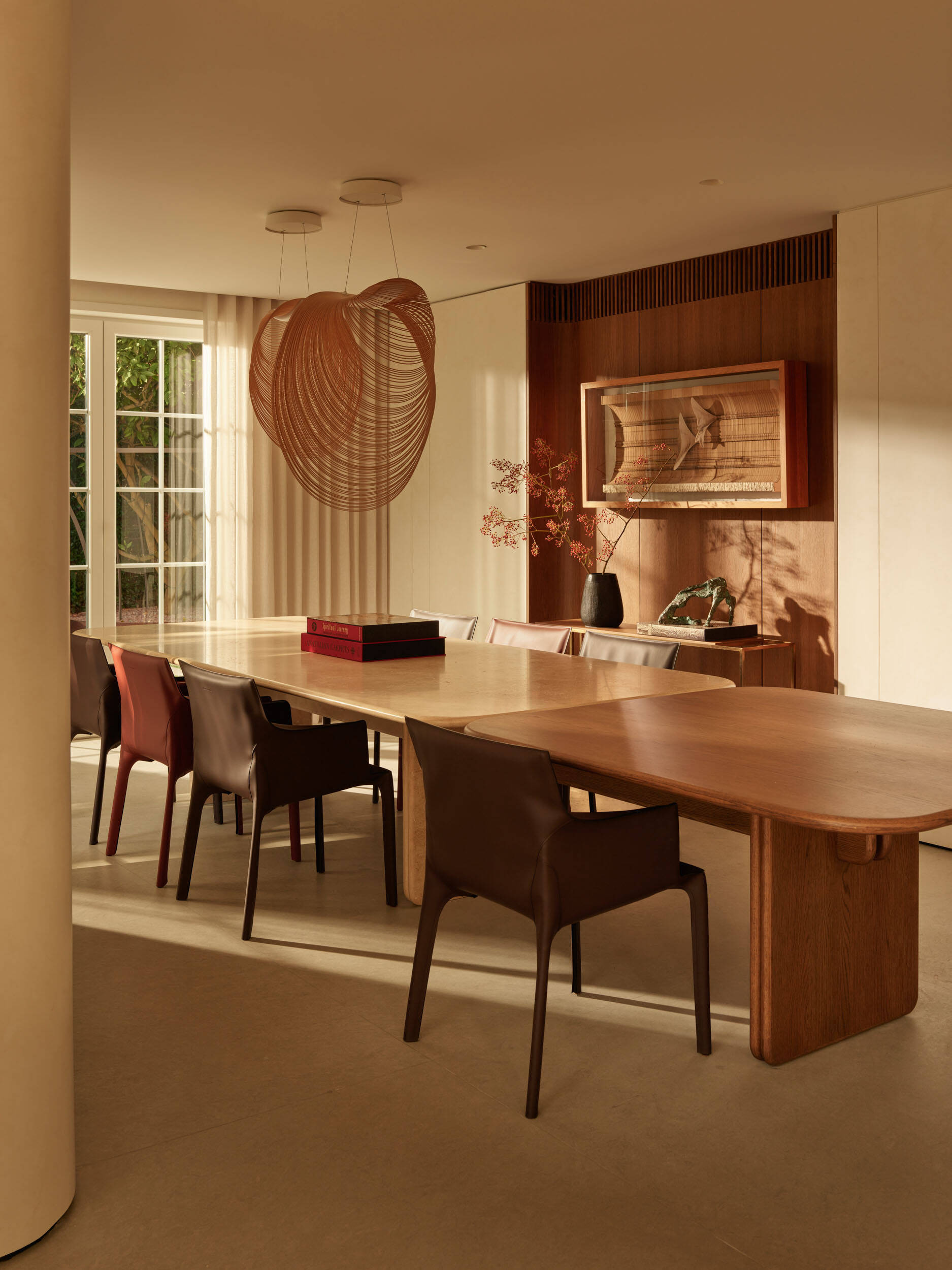 Golden light fills the formal dining room through French doors, highlighting the smooth pale stone and warm wood of the tables. Sculptural chairs in brown and rust tones complement the space, while airy woven pendants add texture and shadow. A wood-backed display niche with simple objects keeps the room polished but approachable for everyday meals.