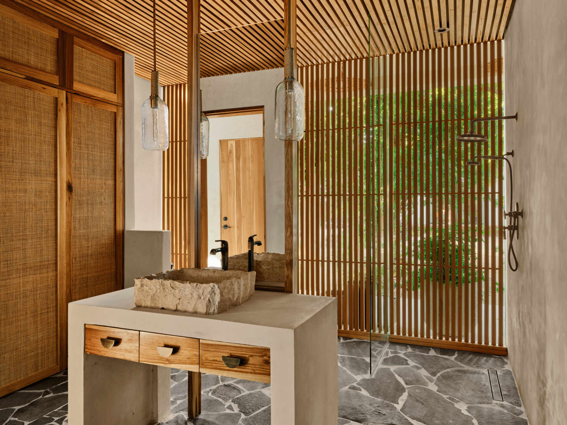 In this bathroom, a carved stone basin sits on a concrete vanity with warm wood drawers, and is paired with simple black fixtures. Vertical timber slats filter light and views toward greenery, while the stone floor and open shower keep the space connected to the outdoors. Textured surfaces and restrained detailing create a bathroom that feels calm, grounded, and closely tied to its tropical setting.