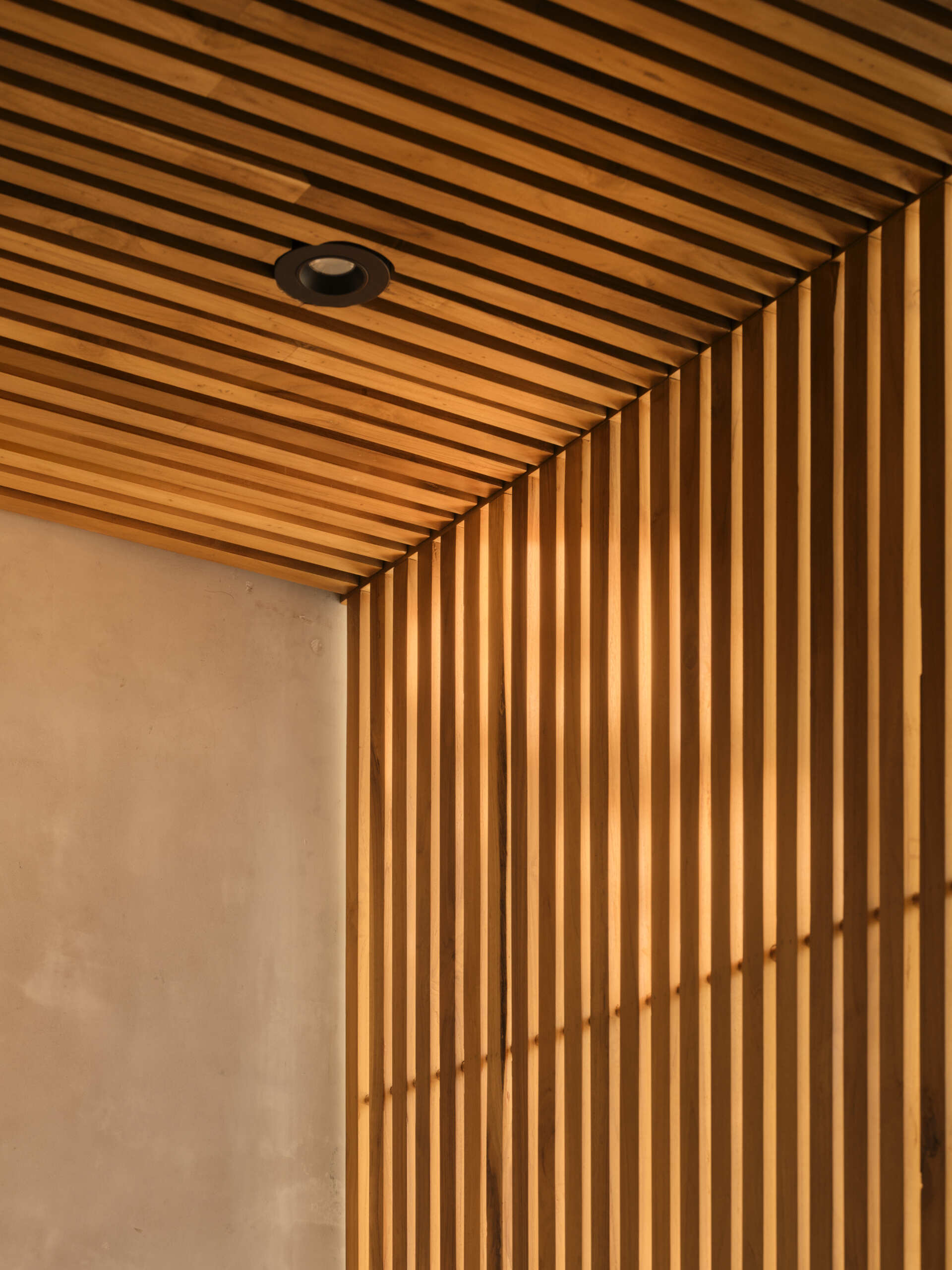 In this bathroom, a carved stone basin sits on a concrete vanity with warm wood drawers, and is paired with simple black fixtures. Vertical timber slats filter light and views toward greenery, while the stone floor and open shower keep the space connected to the outdoors. Textured surfaces and restrained detailing create a bathroom that feels calm, grounded, and closely tied to its tropical setting.