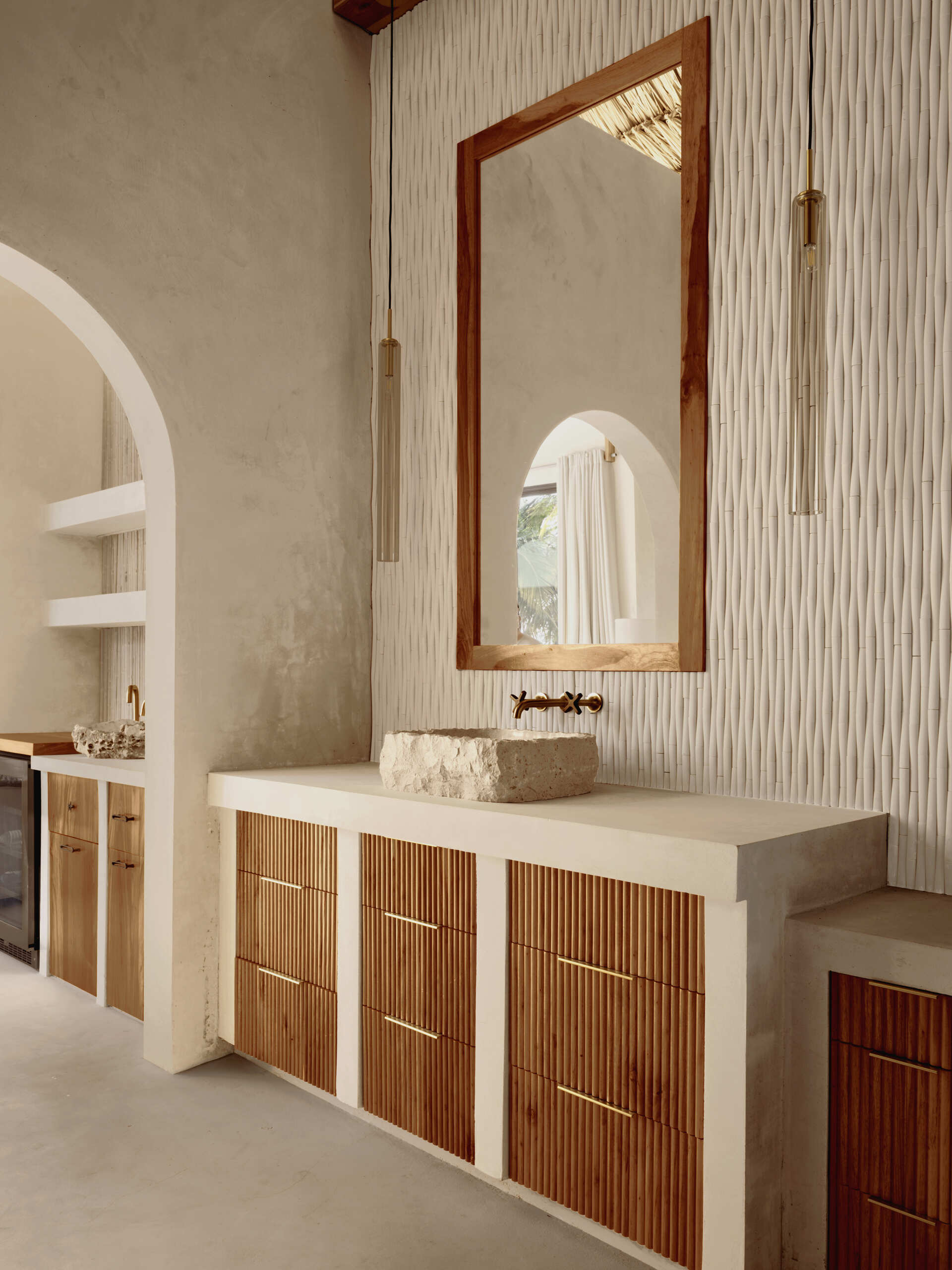 Soft, sandy neutrals and tactile finishes give the bathroom a serene, spa-like feel. Traditional Mayan Chukum plaster adds natural texture and helps regulate temperature, while pale stone walls curve into a sculptural shower, with warm brass fixtures adding a subtle glow. The shower blurs indoor and outdoor living, with a clear glass wall and a door that opens to an outdoor shower featuring rough-cut stone, black pebbles, and matte black fixtures for a calm, resort-like moment at home.