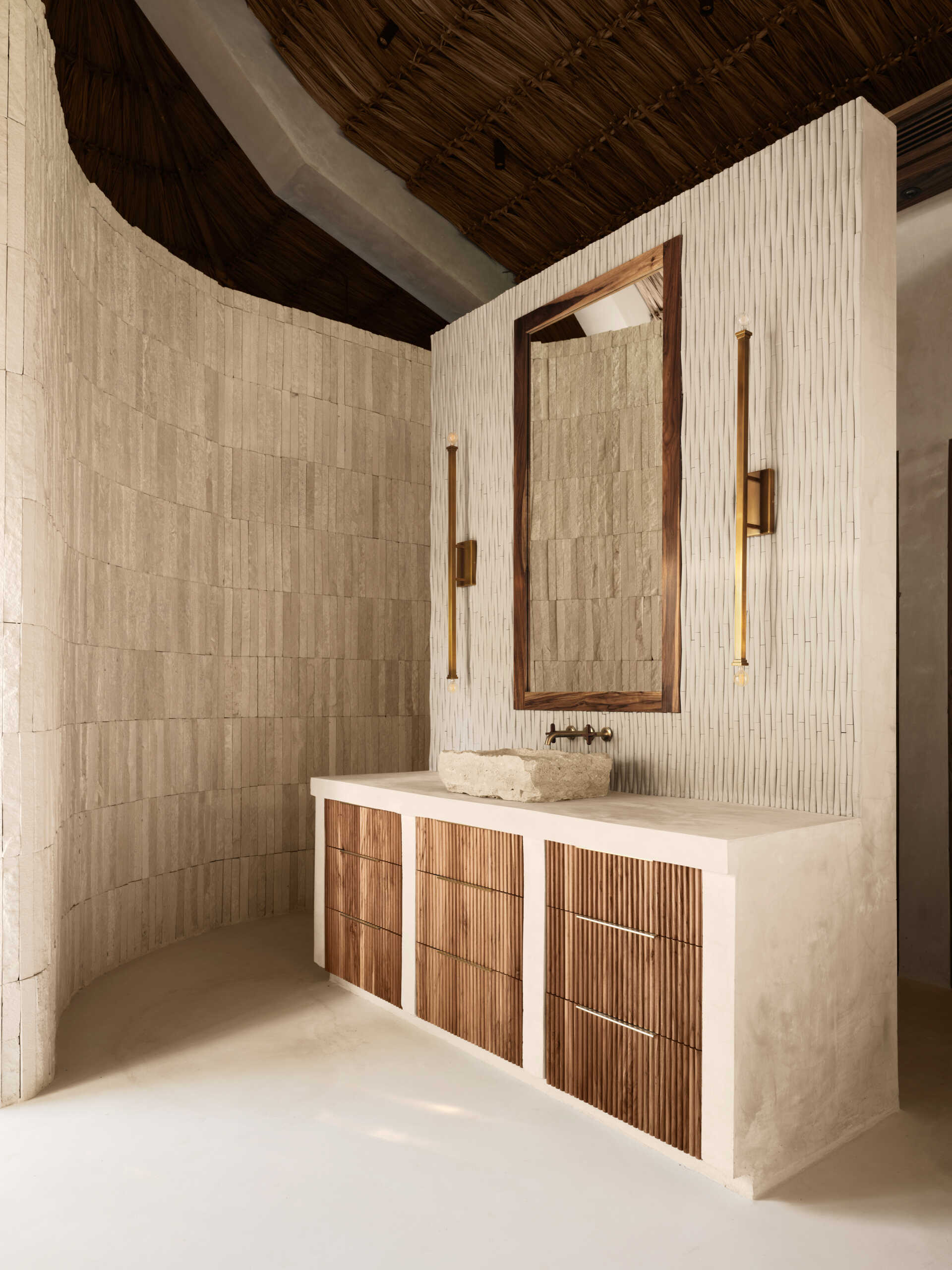 Soft, sandy neutrals and tactile finishes give the bathroom a serene, spa-like feel. Traditional Mayan Chukum plaster adds natural texture and helps regulate temperature, while pale stone walls curve into a sculptural shower, with warm brass fixtures adding a subtle glow. The shower blurs indoor and outdoor living, with a clear glass wall and a door that opens to an outdoor shower featuring rough-cut stone, black pebbles, and matte black fixtures for a calm, resort-like moment at home.