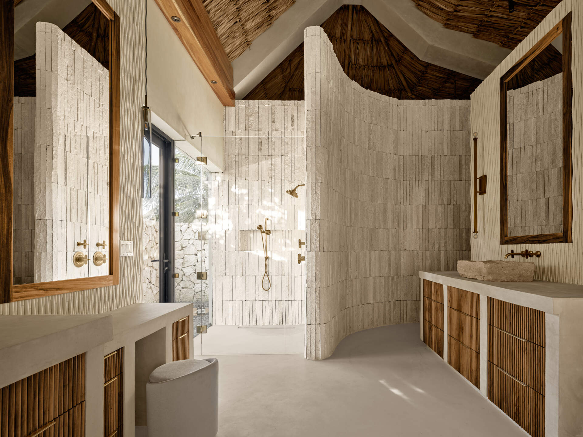 Soft, sandy neutrals and tactile finishes give the bathroom a serene, spa-like feel. Traditional Mayan Chukum plaster adds natural texture and helps regulate temperature, while pale stone walls curve into a sculptural shower, with warm brass fixtures adding a subtle glow. The shower blurs indoor and outdoor living, with a clear glass wall and a door that opens to an outdoor shower featuring rough-cut stone, black pebbles, and matte black fixtures for a calm, resort-like moment at home.