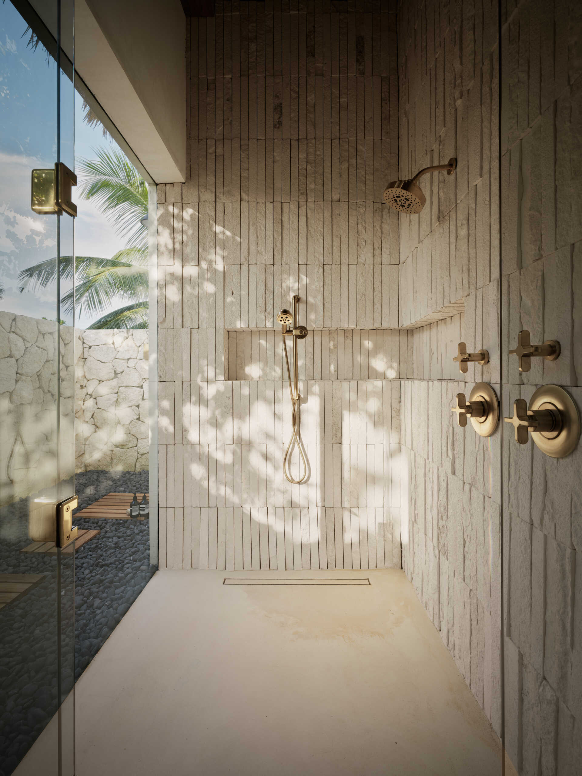 Soft, sandy neutrals and tactile finishes give the bathroom a serene, spa-like feel. Traditional Mayan Chukum plaster adds natural texture and helps regulate temperature, while pale stone walls curve into a sculptural shower, with warm brass fixtures adding a subtle glow. The shower blurs indoor and outdoor living, with a clear glass wall and a door that opens to an outdoor shower featuring rough-cut stone, black pebbles, and matte black fixtures for a calm, resort-like moment at home.