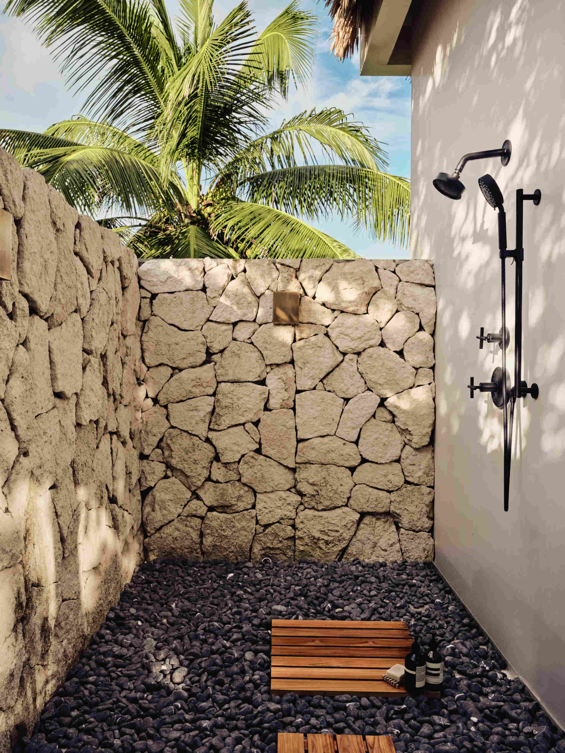 Soft, sandy neutrals and tactile finishes give the bathroom a serene, spa-like feel. Traditional Mayan Chukum plaster adds natural texture and helps regulate temperature, while pale stone walls curve into a sculptural shower, with warm brass fixtures adding a subtle glow. The shower blurs indoor and outdoor living, with a clear glass wall and a door that opens to an outdoor shower featuring rough-cut stone, black pebbles, and matte black fixtures for a calm, resort-like moment at home.