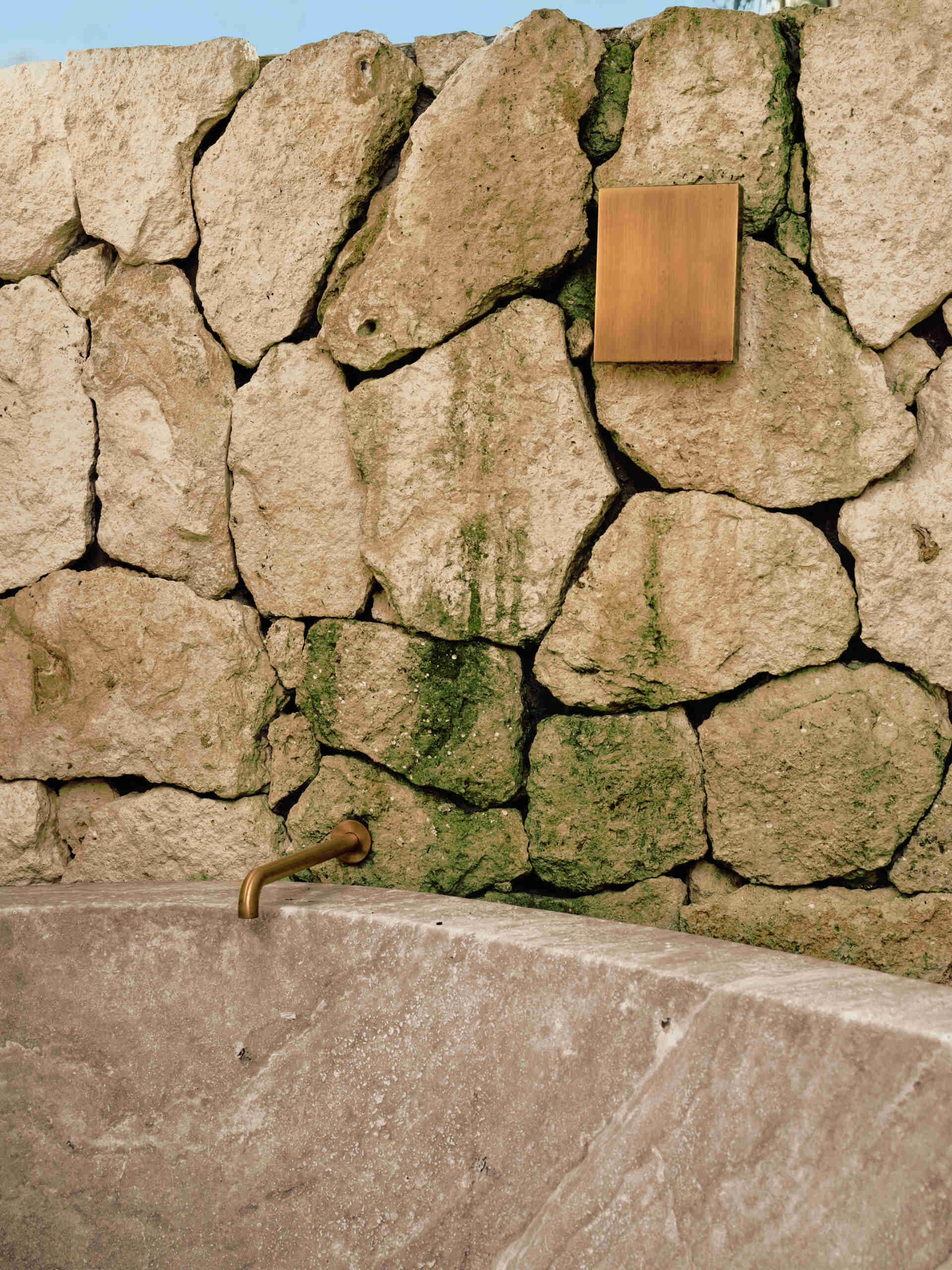 Soft, sandy neutrals and tactile finishes give the bathroom a serene, spa-like feel. Traditional Mayan Chukum plaster adds natural texture and helps regulate temperature, while pale stone walls curve into a sculptural shower, with warm brass fixtures adding a subtle glow. The shower blurs indoor and outdoor living, with a clear glass wall and a door that opens to an outdoor shower featuring rough-cut stone, black pebbles, and matte black fixtures for a calm, resort-like moment at home.