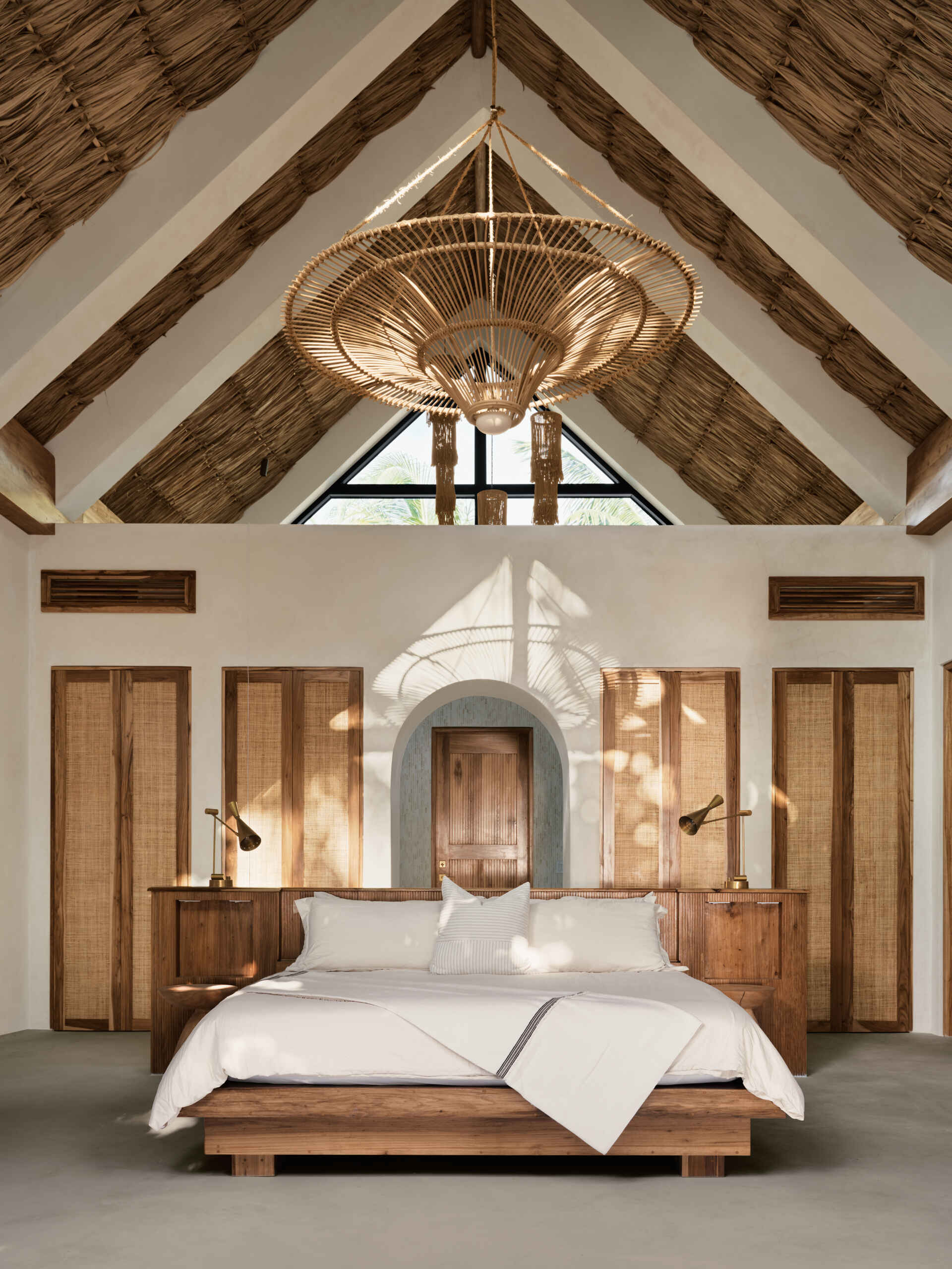 In this bedroom, a low timber bed and integrated wood headboard anchor the space, while soft textiles and neutral tones keep the atmosphere relaxed. Above, thatched ceiling and exposed beams add texture and warmth, reinforcing the connection to local craft. Large sliding doors open the rooms to surrounding greenery, allowing light and breezes to shape the experience throughout the day.