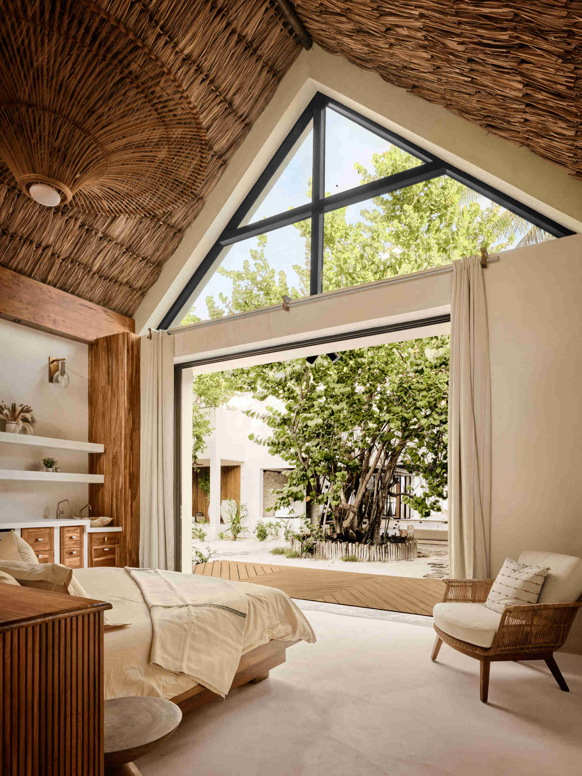 In this bedroom, a low timber bed and integrated wood headboard anchor the space, while soft textiles and neutral tones keep the atmosphere relaxed. Above, thatched ceiling and exposed beams add texture and warmth, reinforcing the connection to local craft. Large sliding doors open the rooms to surrounding greenery, allowing light and breezes to shape the experience throughout the day.