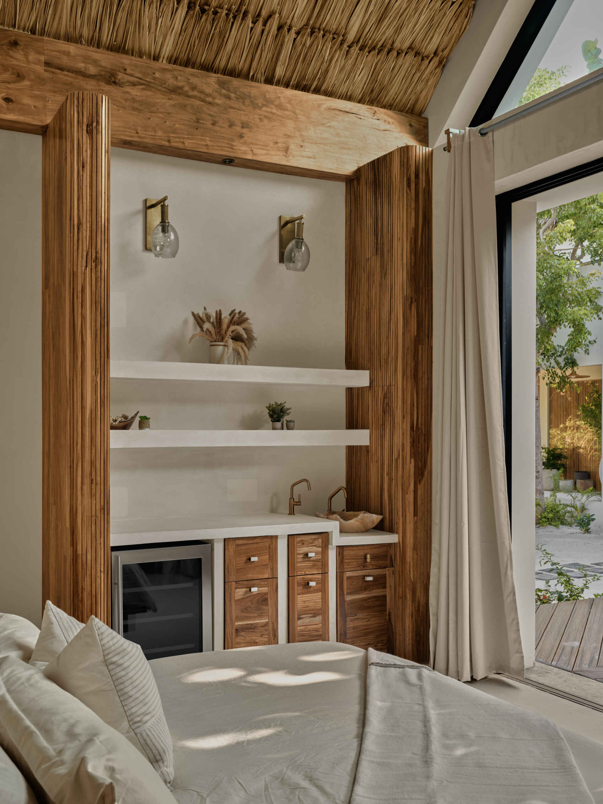In this bedroom, a low timber bed and integrated wood headboard anchor the space, while soft textiles and neutral tones keep the atmosphere relaxed. Above, thatched ceiling and exposed beams add texture and warmth, reinforcing the connection to local craft. Large sliding doors open the rooms to surrounding greenery, allowing light and breezes to shape the experience throughout the day.