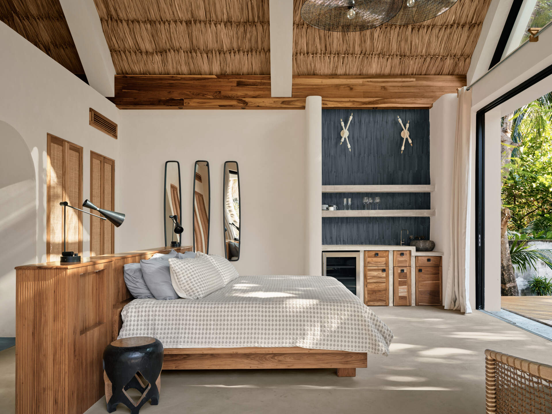 In this bedroom, a low timber bed and integrated wood headboard anchor the space, while soft textiles and neutral tones keep the atmosphere relaxed. Above, thatched ceiling and exposed beams add texture and warmth, reinforcing the connection to local craft. Large sliding doors open the rooms to surrounding greenery, allowing light and breezes to shape the experience throughout the day.