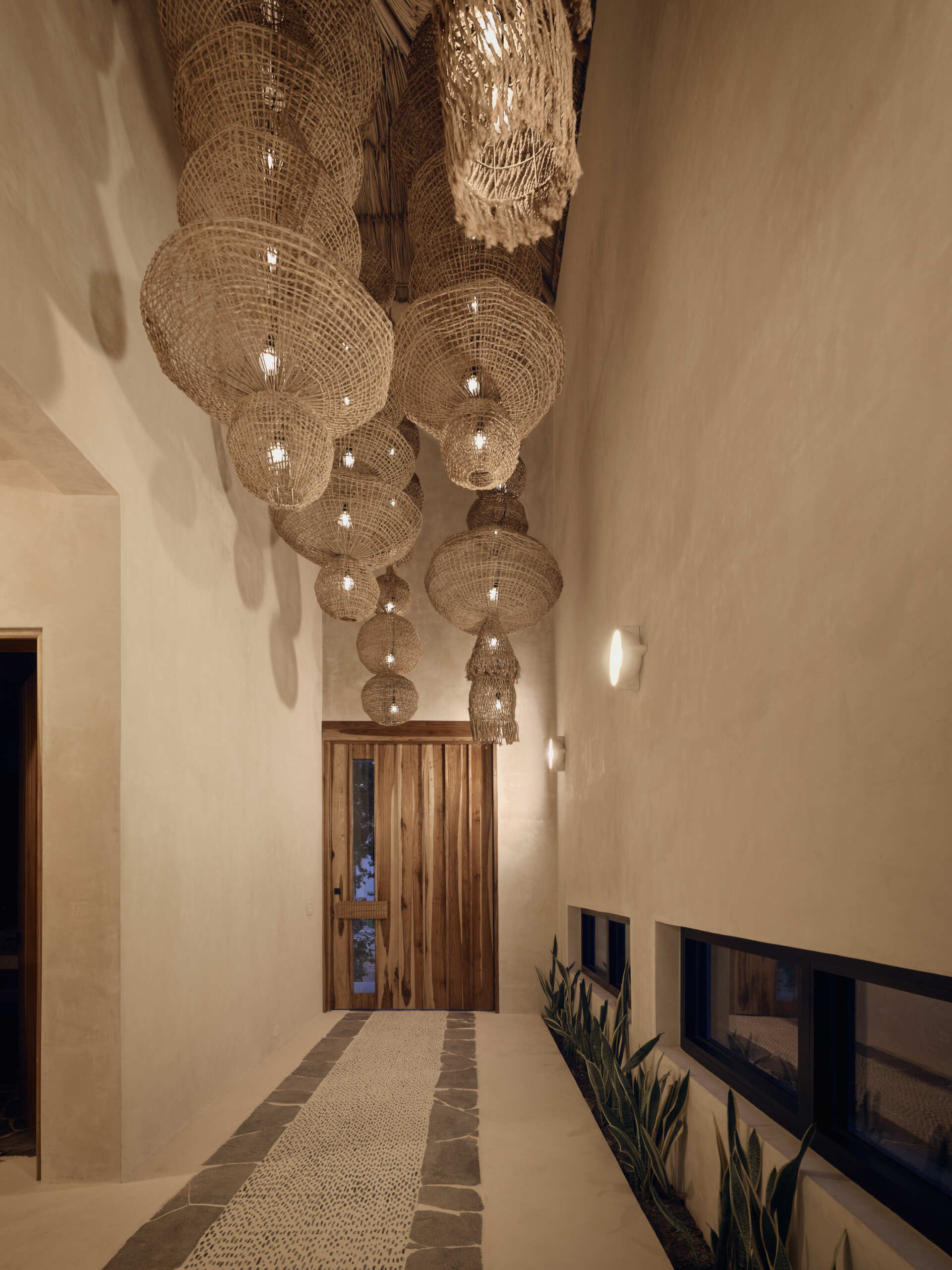 The entryway sets a calm, tactile tone from the moment you step inside. Soft Chukum plaster walls rise to a high ceiling where a cluster of handcrafted woven pendants creates a warm, sculptural canopy of light. Underfoot, a textured stone and pebble path draws the eye toward the solid wood front door, while low narrow windows and planted greenery along the wall keep the space grounded and cool.