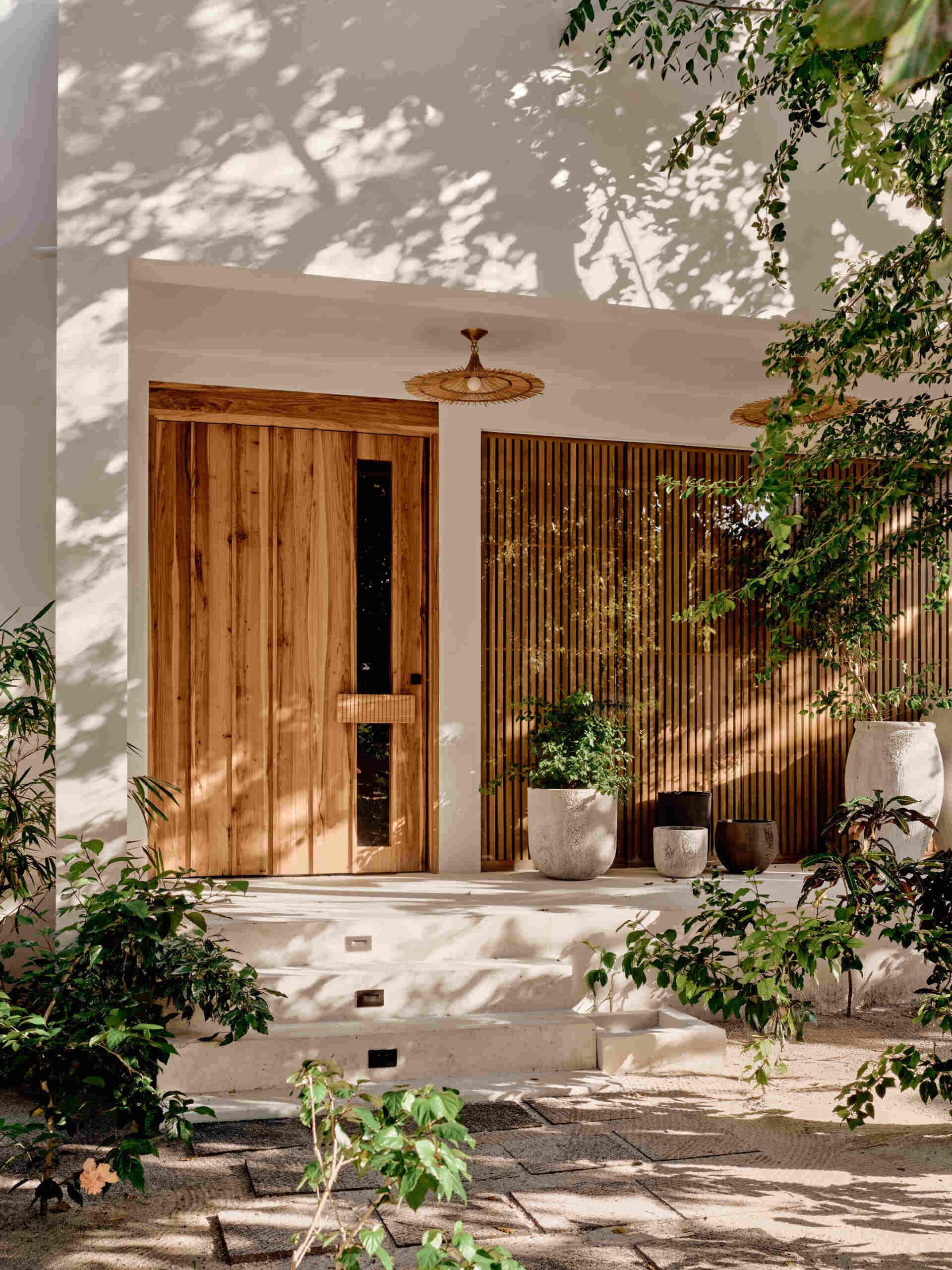 A solid wood door welcomes visitors to the home and sits within smooth pale walls, which are paired with vertical wood screening that filters views and casts shifting shadows throughout the day.