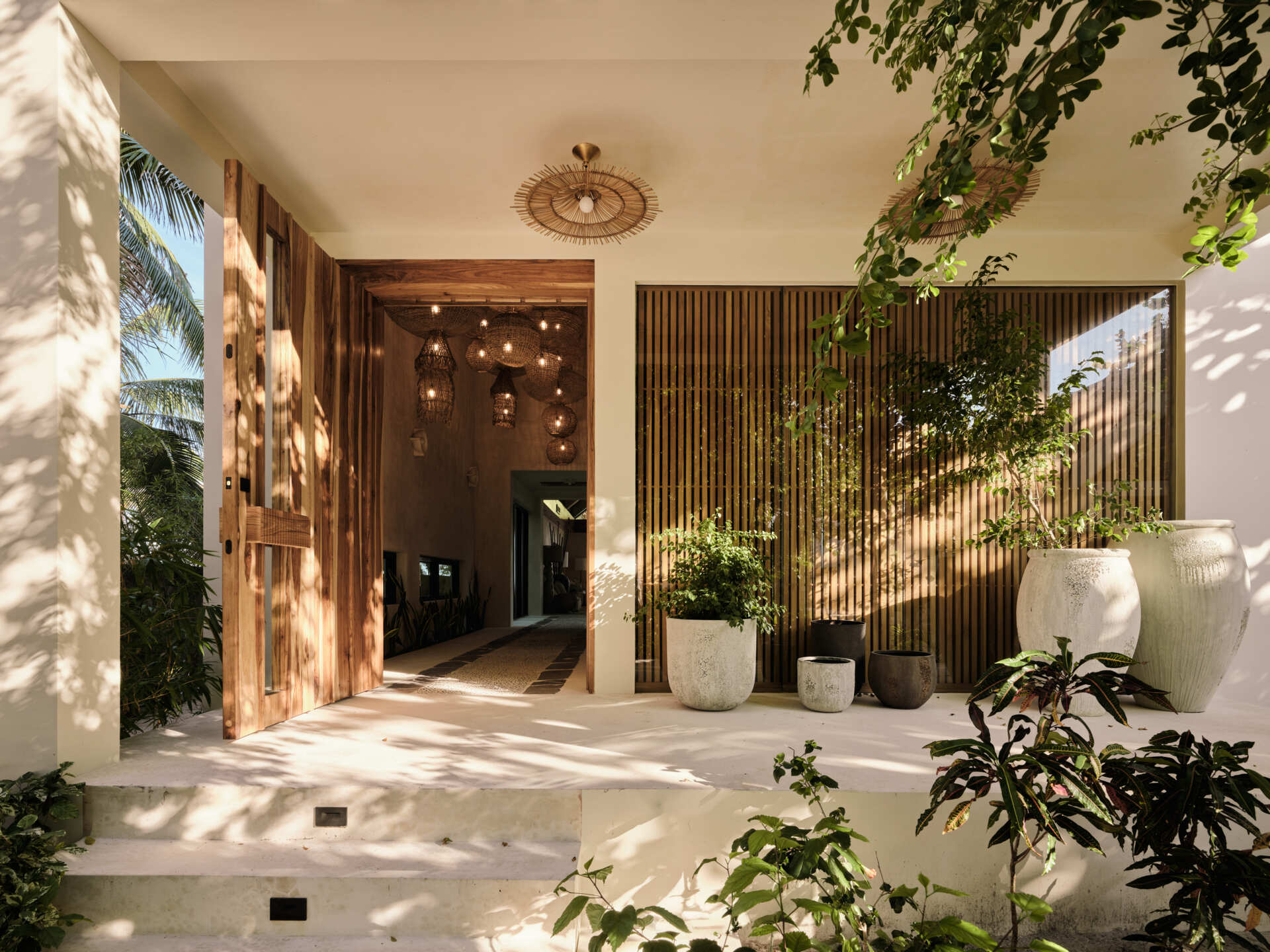 A solid wood door welcomes visitors to the home and sits within smooth pale walls, which are paired with vertical wood screening that filters views and casts shifting shadows throughout the day.