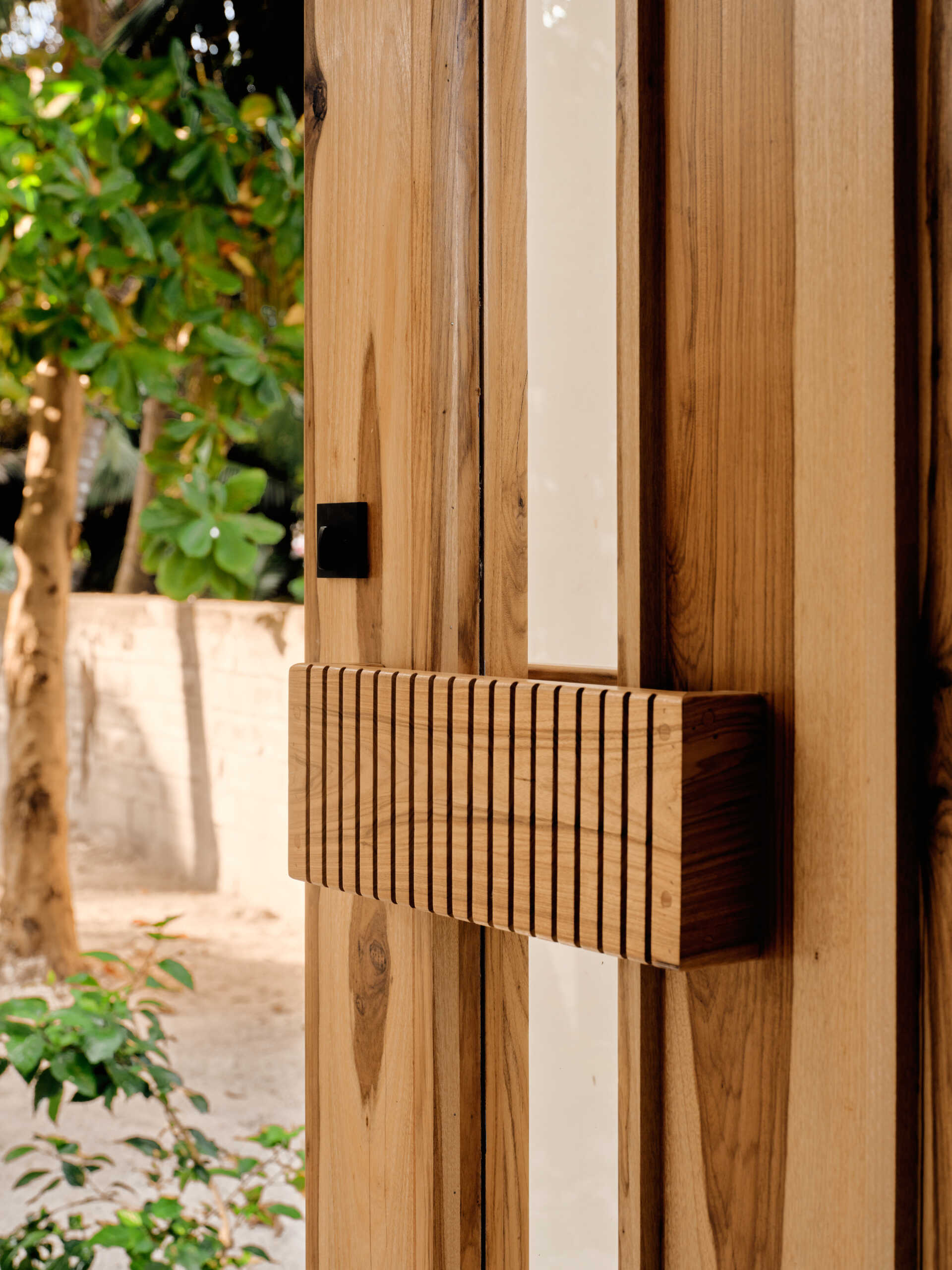 A solid wood door welcomes visitors to the home and sits within smooth pale walls, which are paired with vertical wood screening that filters views and casts shifting shadows throughout the day.
