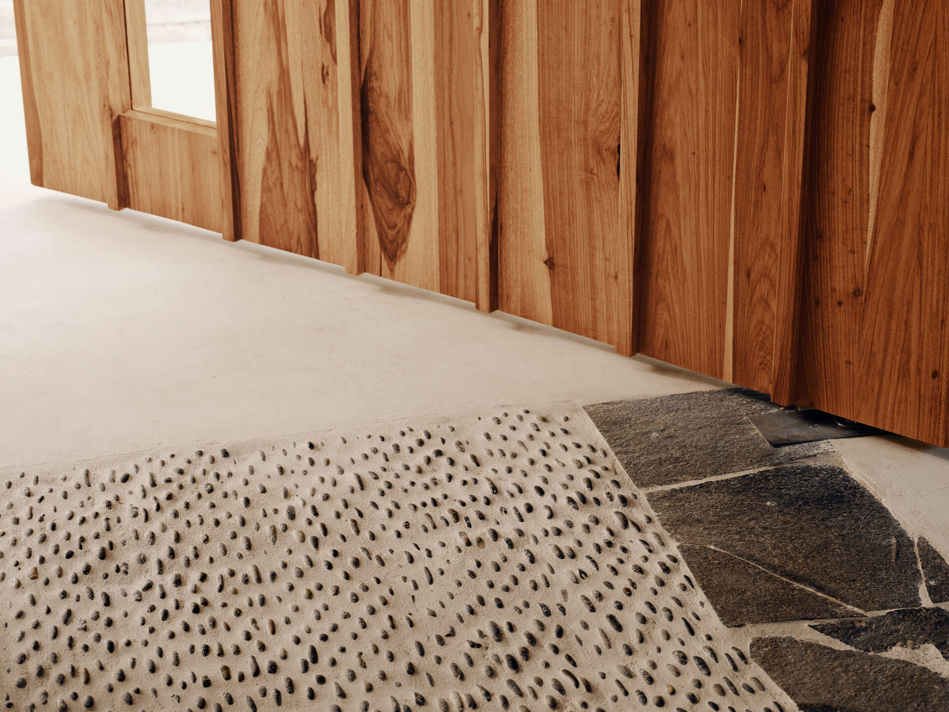 A solid wood door welcomes visitors to the home and sits within smooth pale walls, which are paired with vertical wood screening that filters views and casts shifting shadows throughout the day.