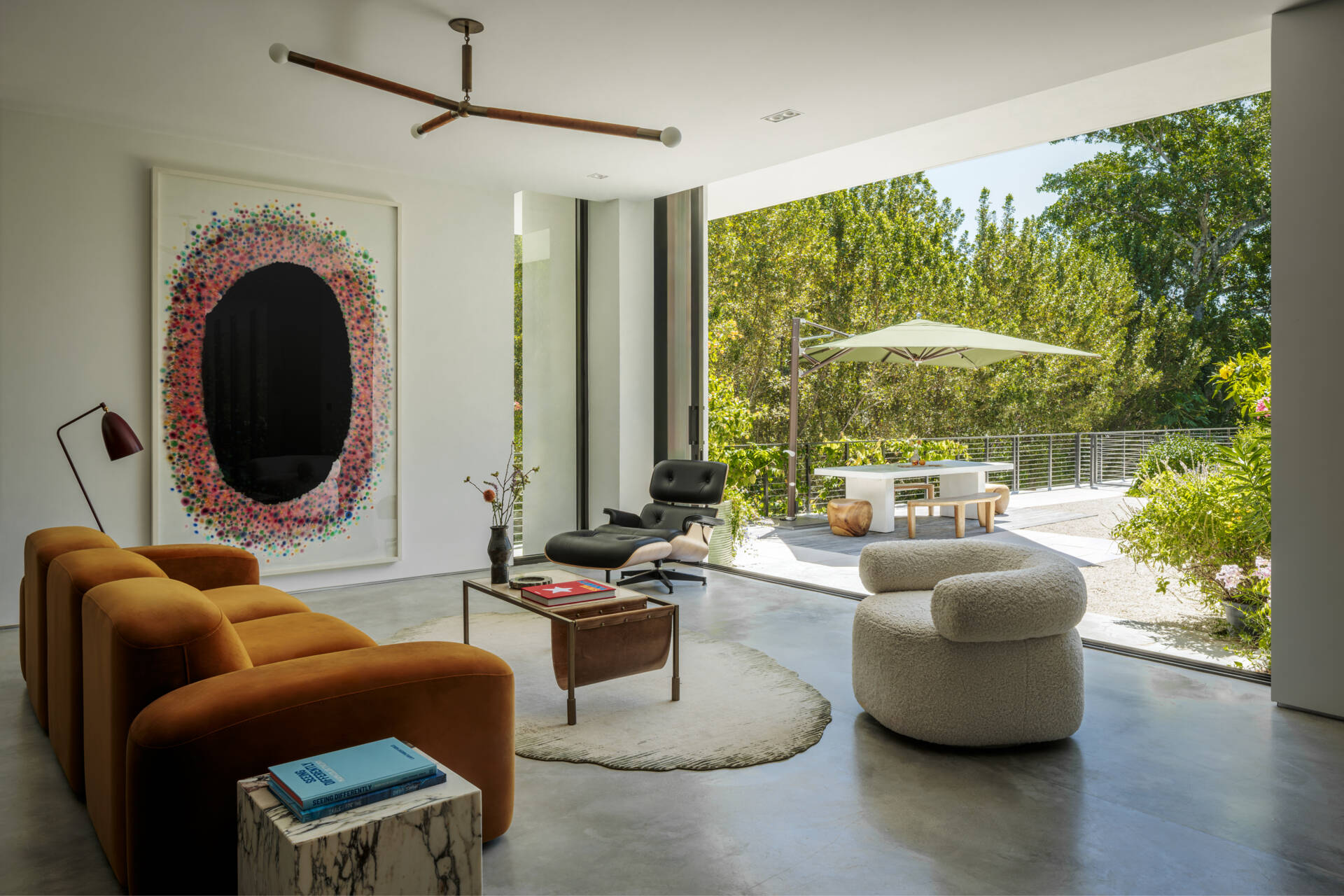 This modern living space opens seamlessly to the patio, turning outdoor areas into part of the room. A caramel velvet sofa and boucle chair add warmth and softness, while an oversized artwork adds color, and the shaded outdoor area invites relaxed meals and casual lounging surrounded by greenery.