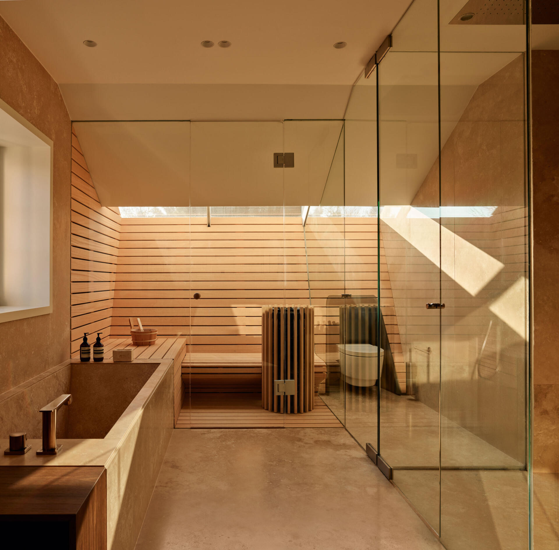 The sauna combines pale wood slats and daylight from above to create a spa-like atmosphere. Smooth stone surfaces and a deep soaking tub keep the palette calm. A glass shower enclosure expands the sense of space, and every detail feels intentional for a restorative experience.