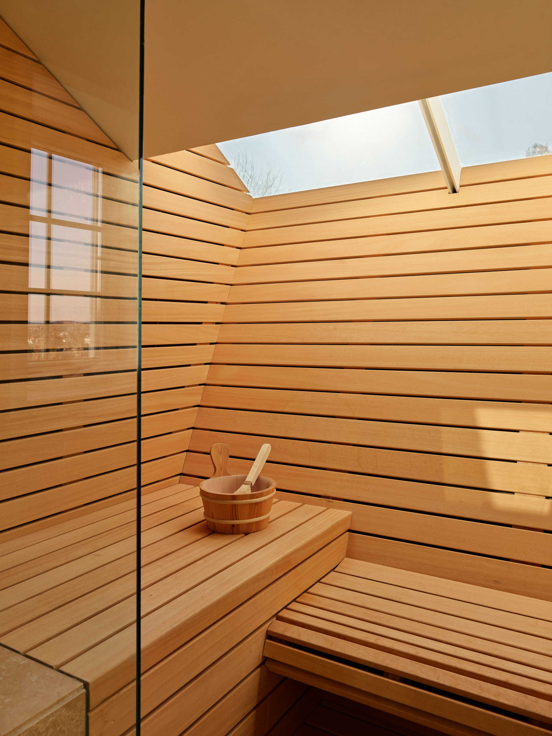 The sauna combines pale wood slats and daylight from above to create a spa-like atmosphere. Smooth stone surfaces and a deep soaking tub keep the palette calm. A glass shower enclosure expands the sense of space, and every detail feels intentional for a restorative experience.