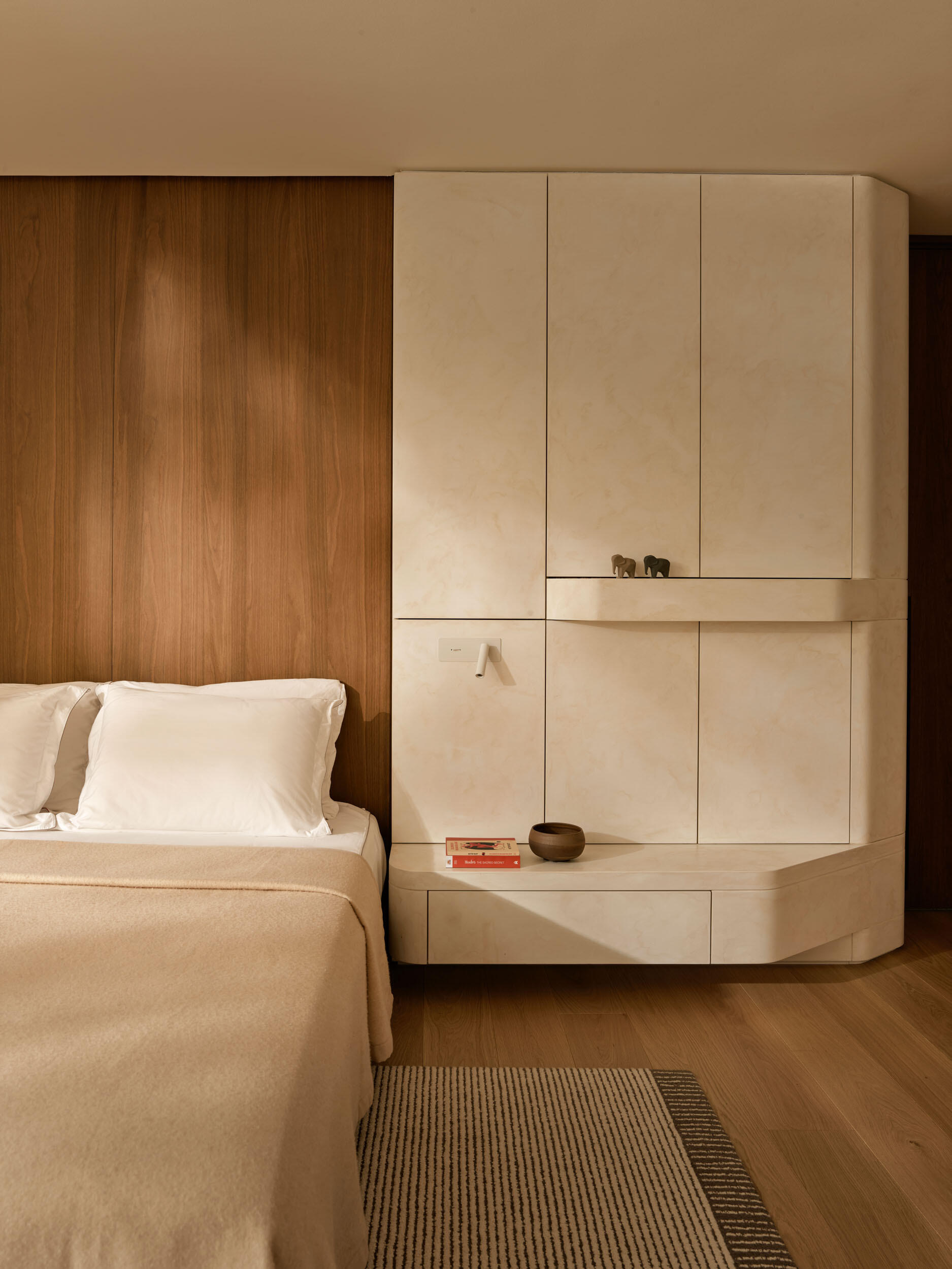 The bedroom is quietly refined, with a warm wood accent wall. Rounded cabinetry provides seamless storage, and gentle light warms the pale surfaces. The ensuite feels like a private spa, with creamy stone surfaces, a built-in tub, and a sleek glass enclosure that lets light travel freely, creating a serene daily experience.