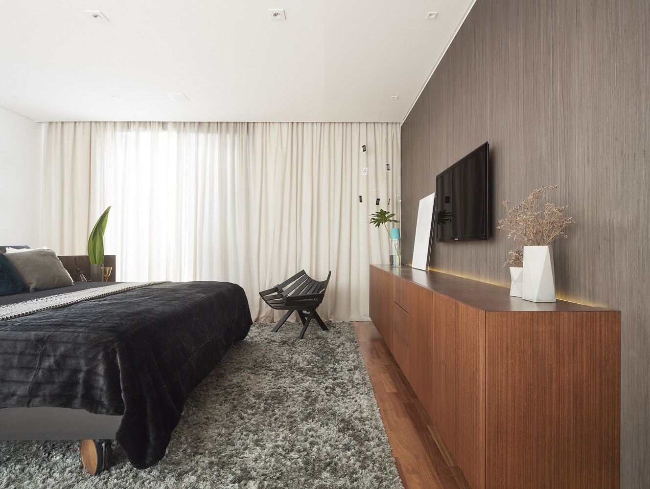 Inside the bedroom, the layout breaks from convention. The bed is positioned at the center of the room, anchored by a curved wood headboard that softens the geometry of the space. Round mirrors reflect sunlight across the room, amplifying natural light and drawing attention to the cabinetry along the opposite wall.