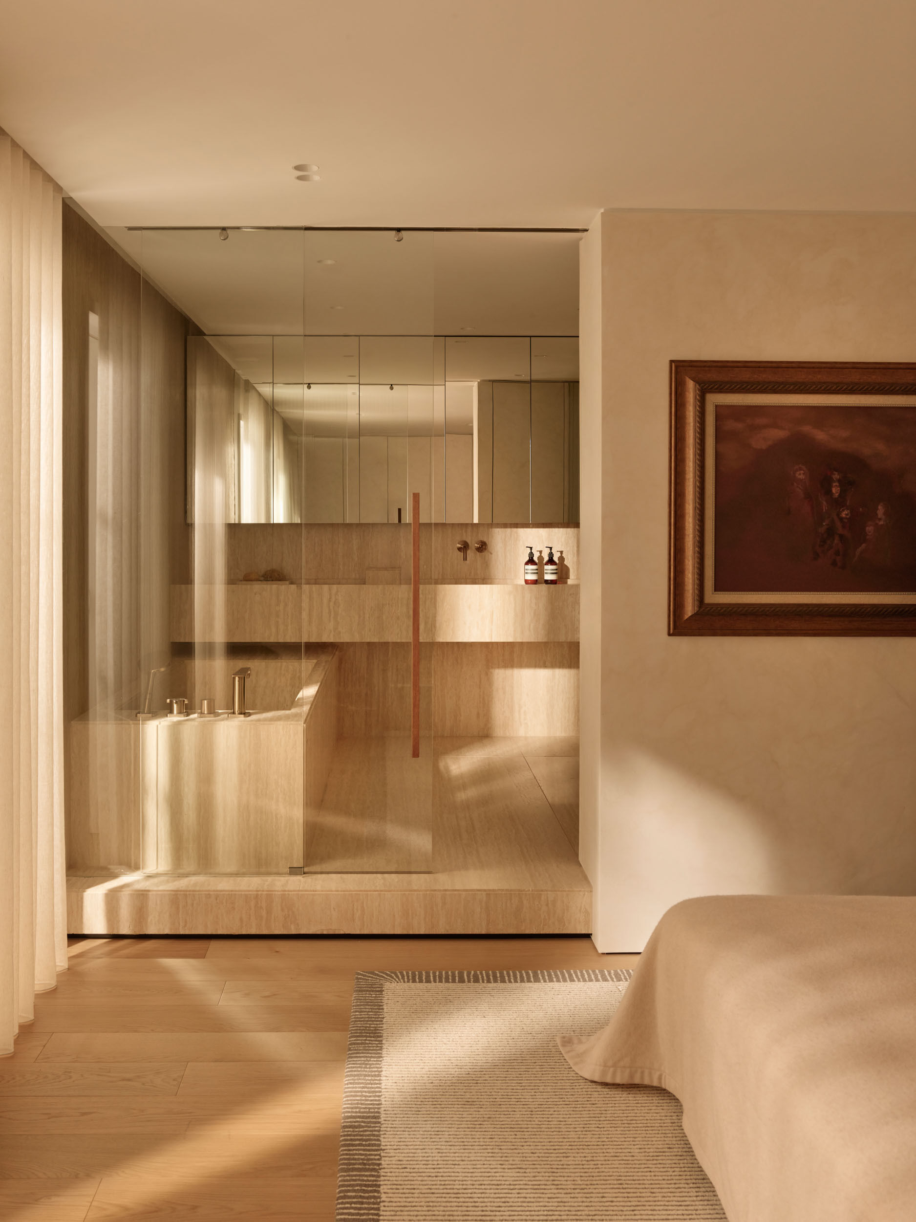 The bedroom is quietly refined, with a warm wood accent wall. Rounded cabinetry provides seamless storage, and gentle light warms the pale surfaces. The ensuite feels like a private spa, with creamy stone surfaces, a built-in tub, and a sleek glass enclosure that lets light travel freely, creating a serene daily experience.